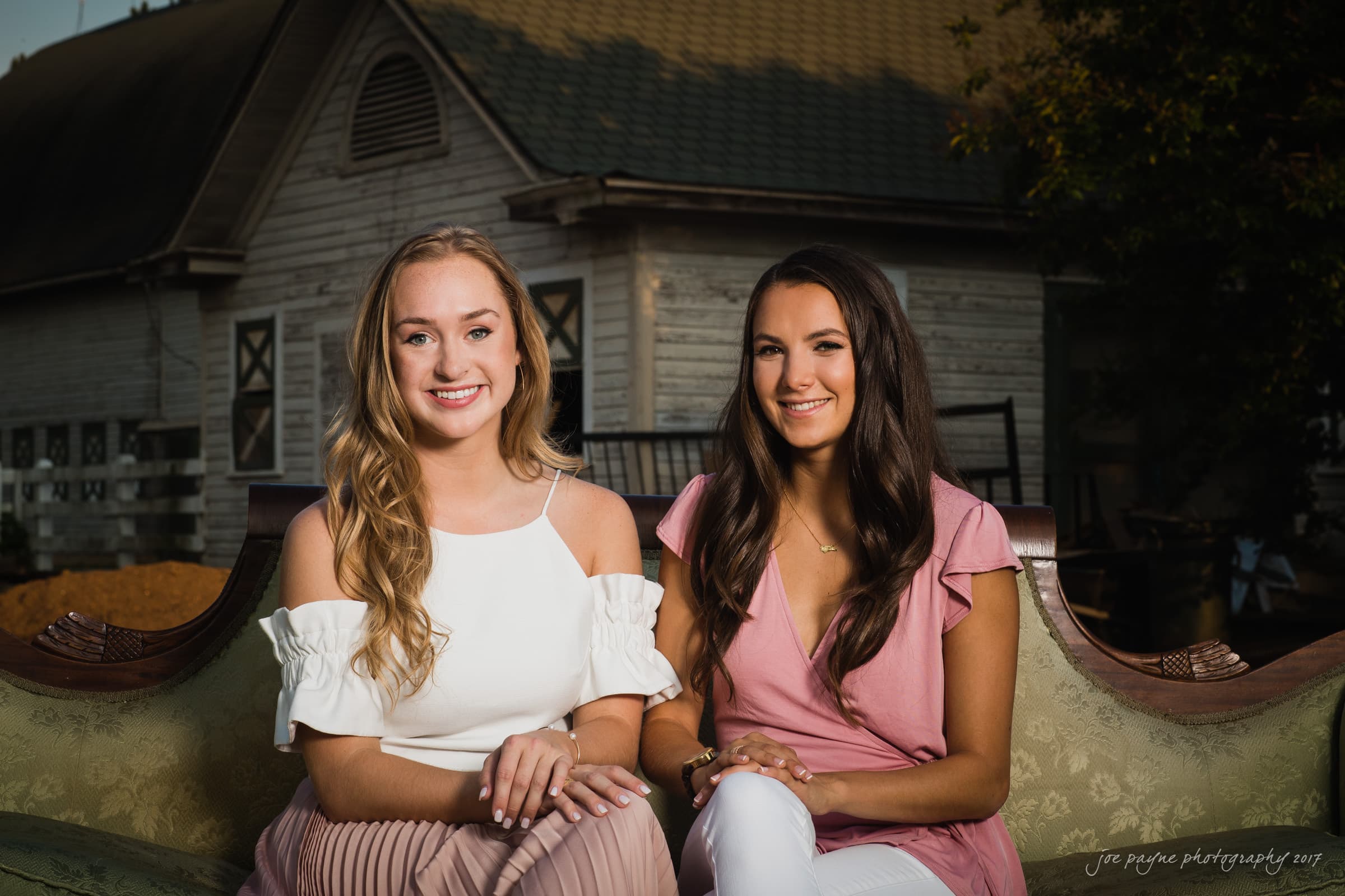 Two women are sitting on an outdoor couch, smiling at the camera. One is wearing a white top, and the other is in a pink outfit. A rustic barn-like building is visible in the background.