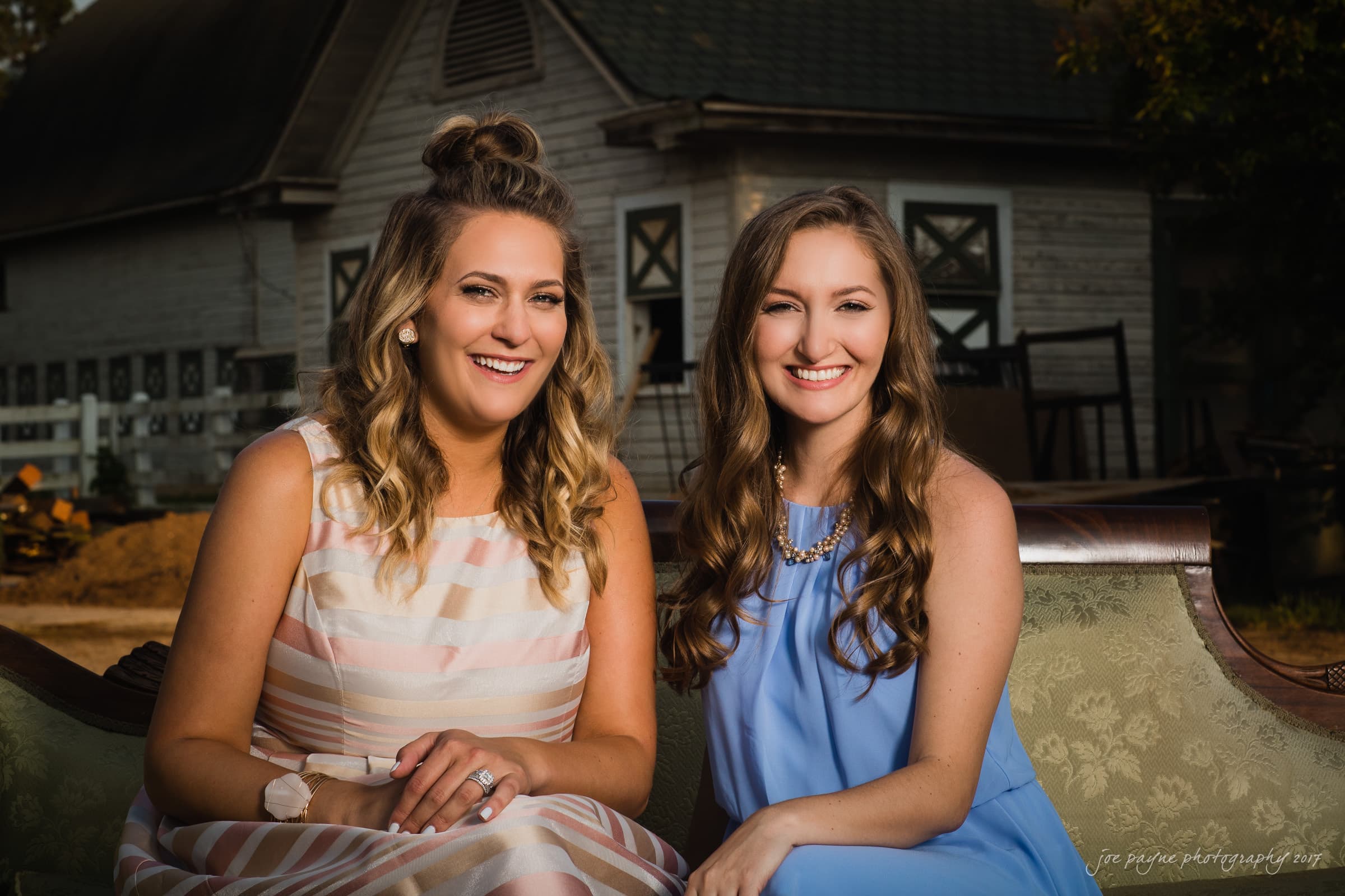Two women are sitting on a vintage sofa outdoors, smiling at the camera. They are both wearing dresses, one in pastel stripes and the other in light blue. A rustic building is in the background.
