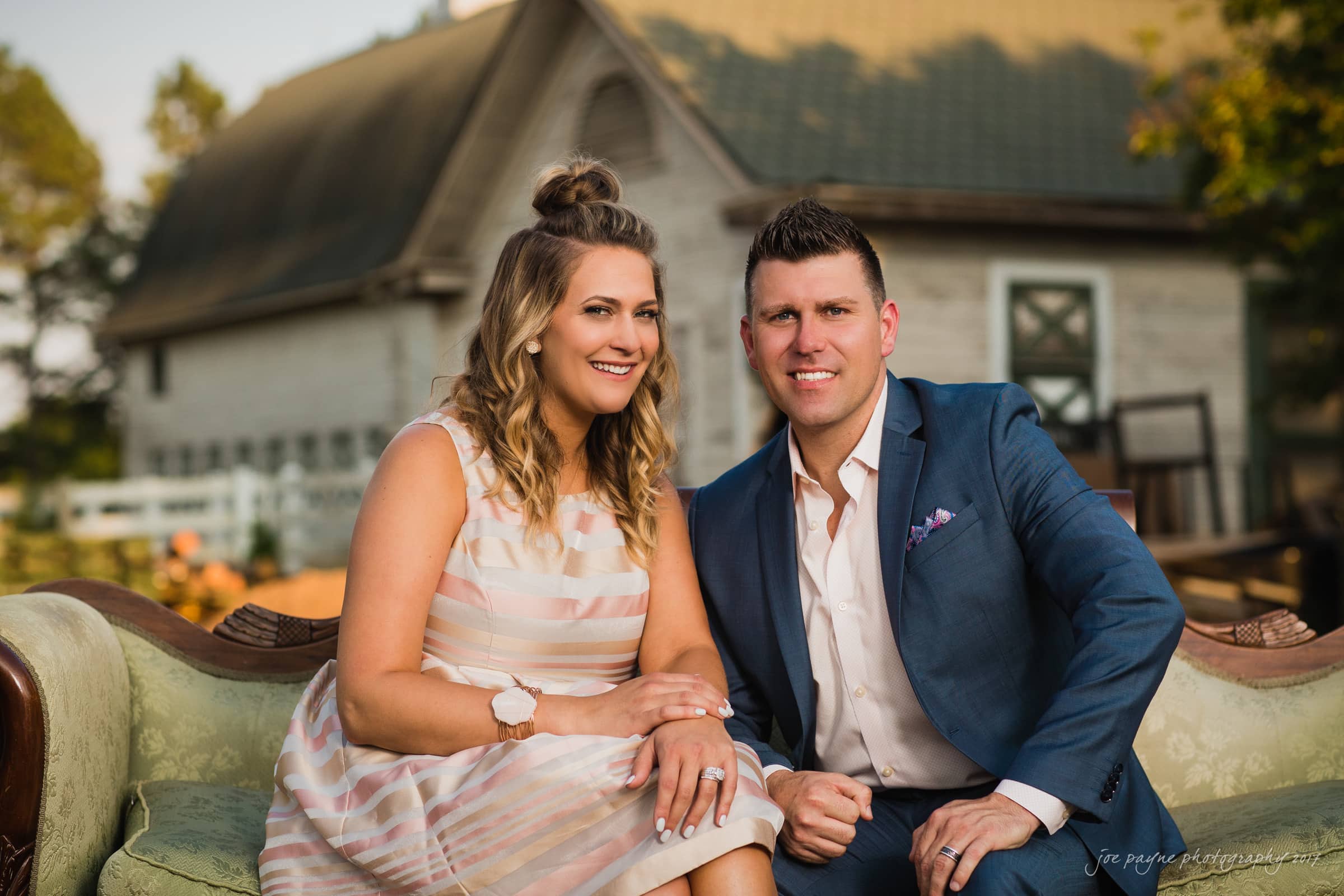 A woman in a striped dress and a man in a blue suit sit together on a vintage couch outdoors. A rustic building is in the background, and both are smiling at the camera.