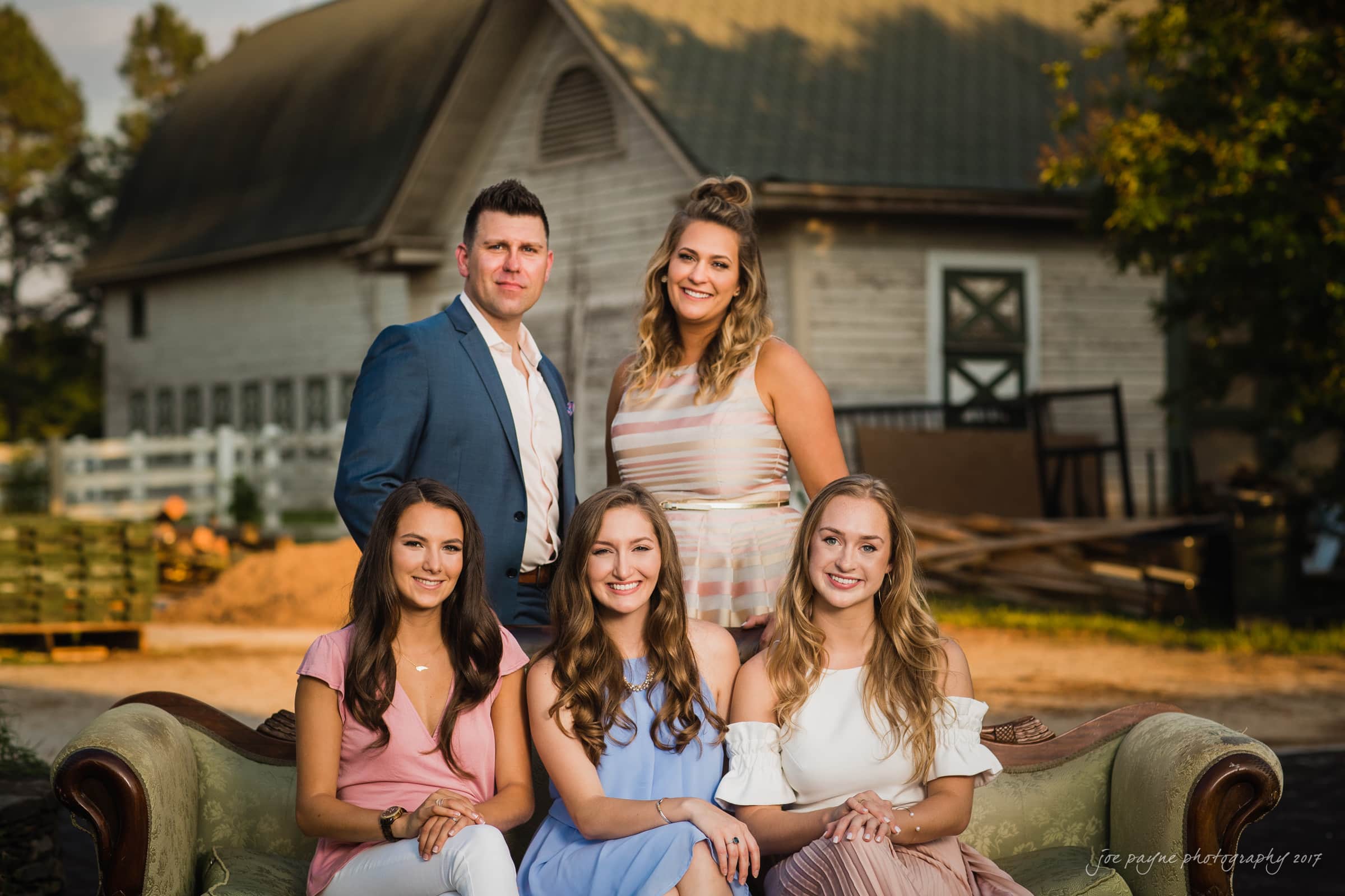 A group of five people, four women and one man, sit and stand around a vintage couch outdoors. They are dressed in semi-formal attire. A rustic barn is in the background under soft, golden-hour lighting.