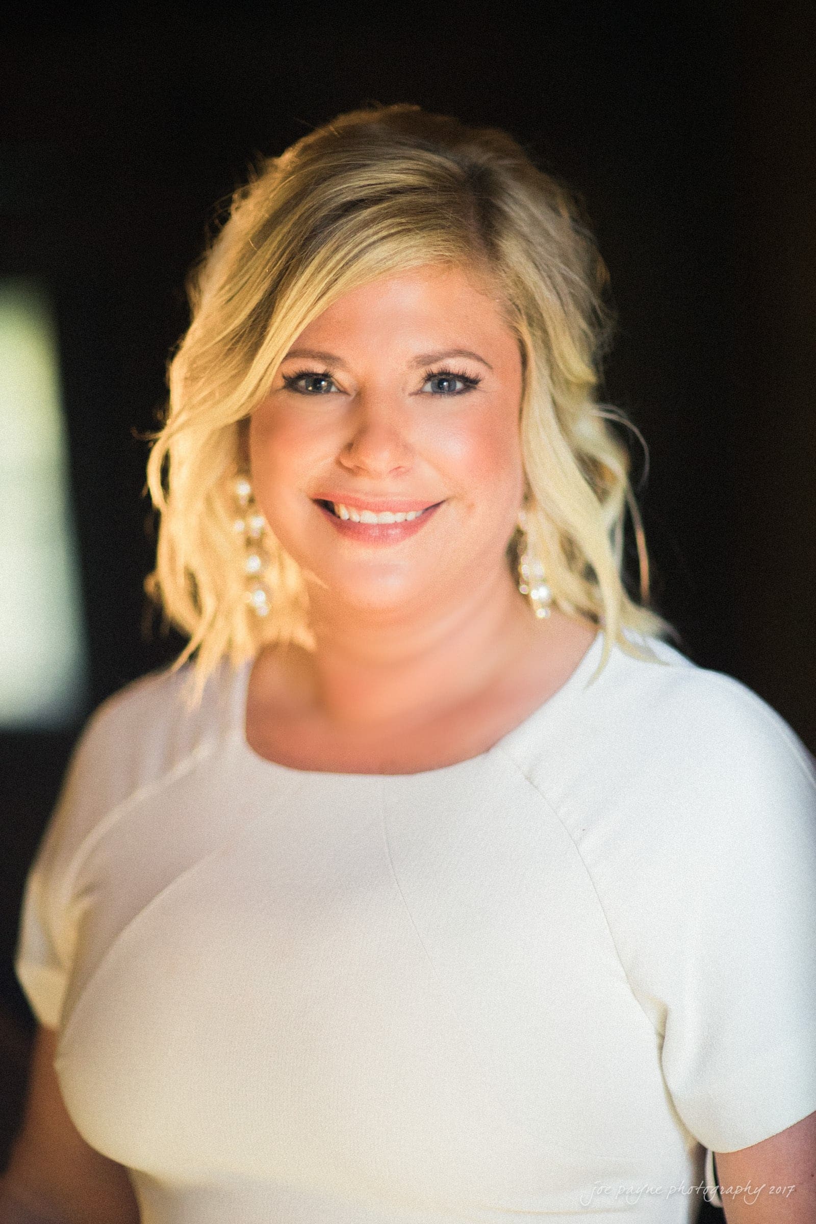 A woman with blond hair smiles while wearing a white top and long, dangly earrings. She stands in a softly lit setting with a dark background.