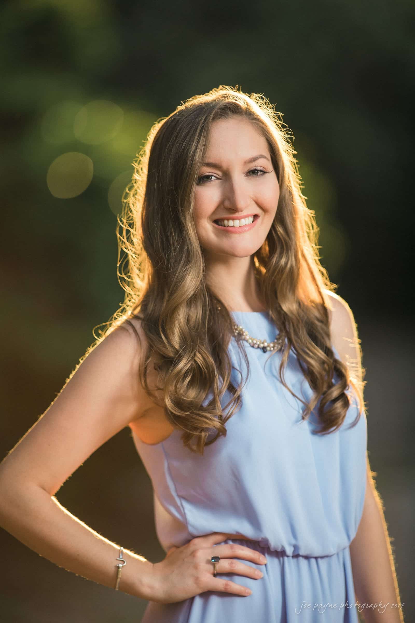 A woman with long wavy hair smiles while standing outdoors. She is wearing a light purple sleeveless dress and a necklace. The background is softly blurred with warm lighting.