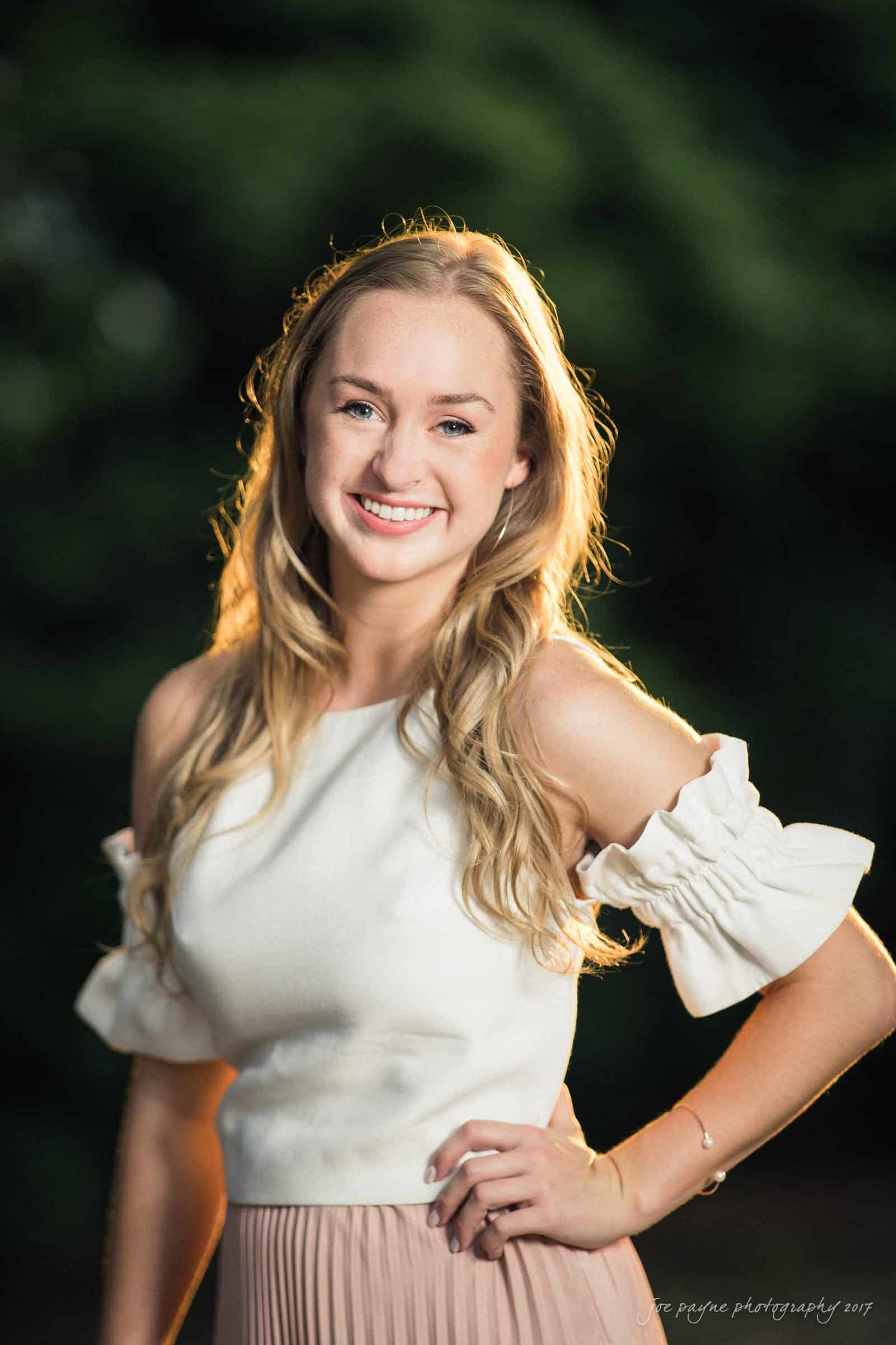 A young woman with long blonde hair smiles at the camera. She is wearing an off-shoulder white top and a pink skirt. The background is blurred with greenery, and there is soft lighting.