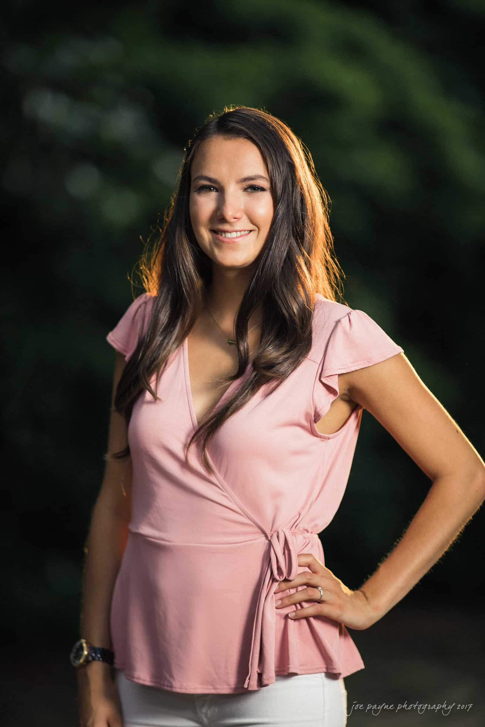 A woman with long brown hair smiles while standing outdoors. She is wearing a pink top and white pants. The background is a blurred green foliage.