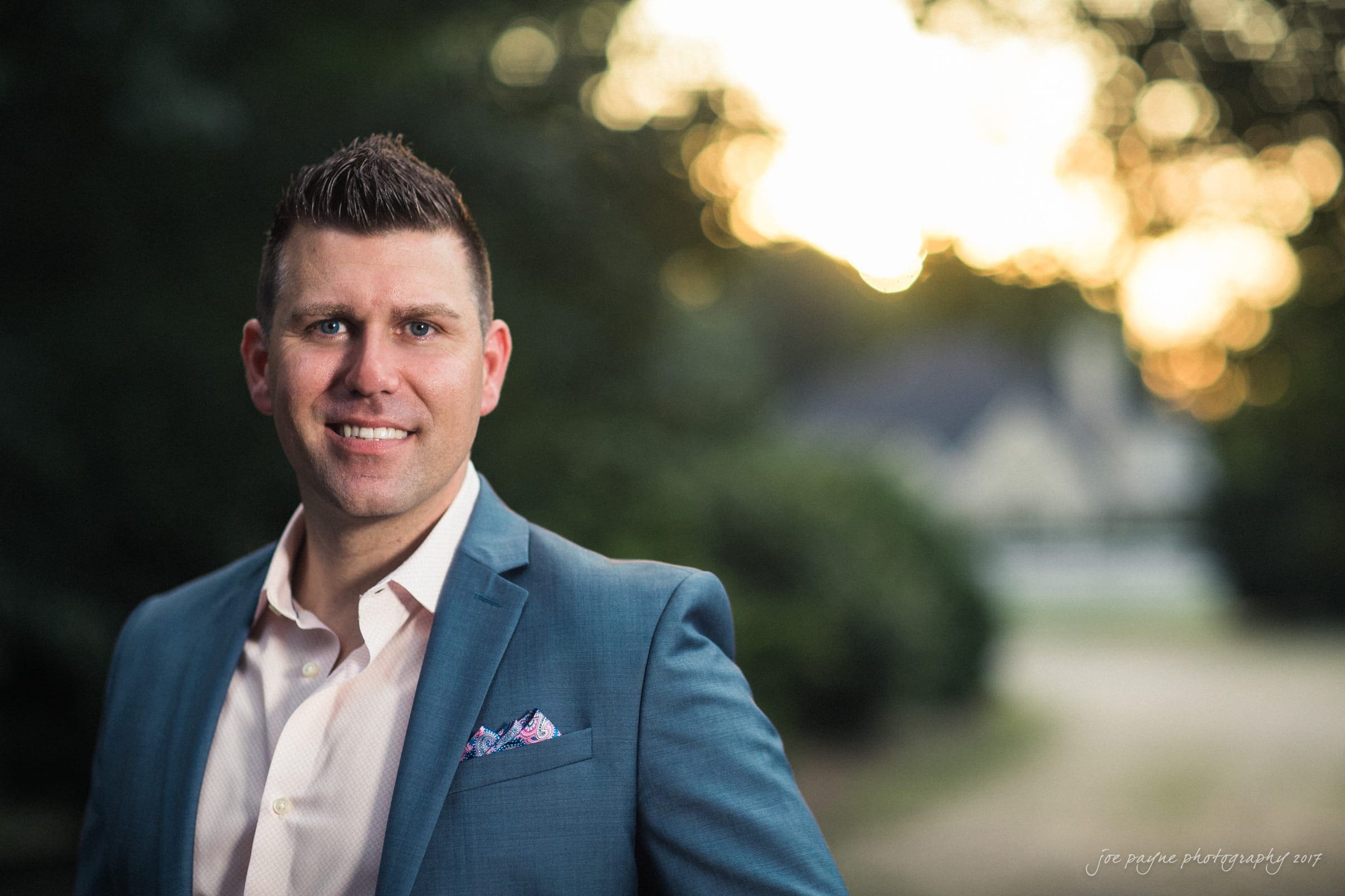 A man in a blue suit and white shirt stands outdoors, smiling at the camera. The background is blurred with greenery and a house visible, and the sun is setting, creating a warm glow.
