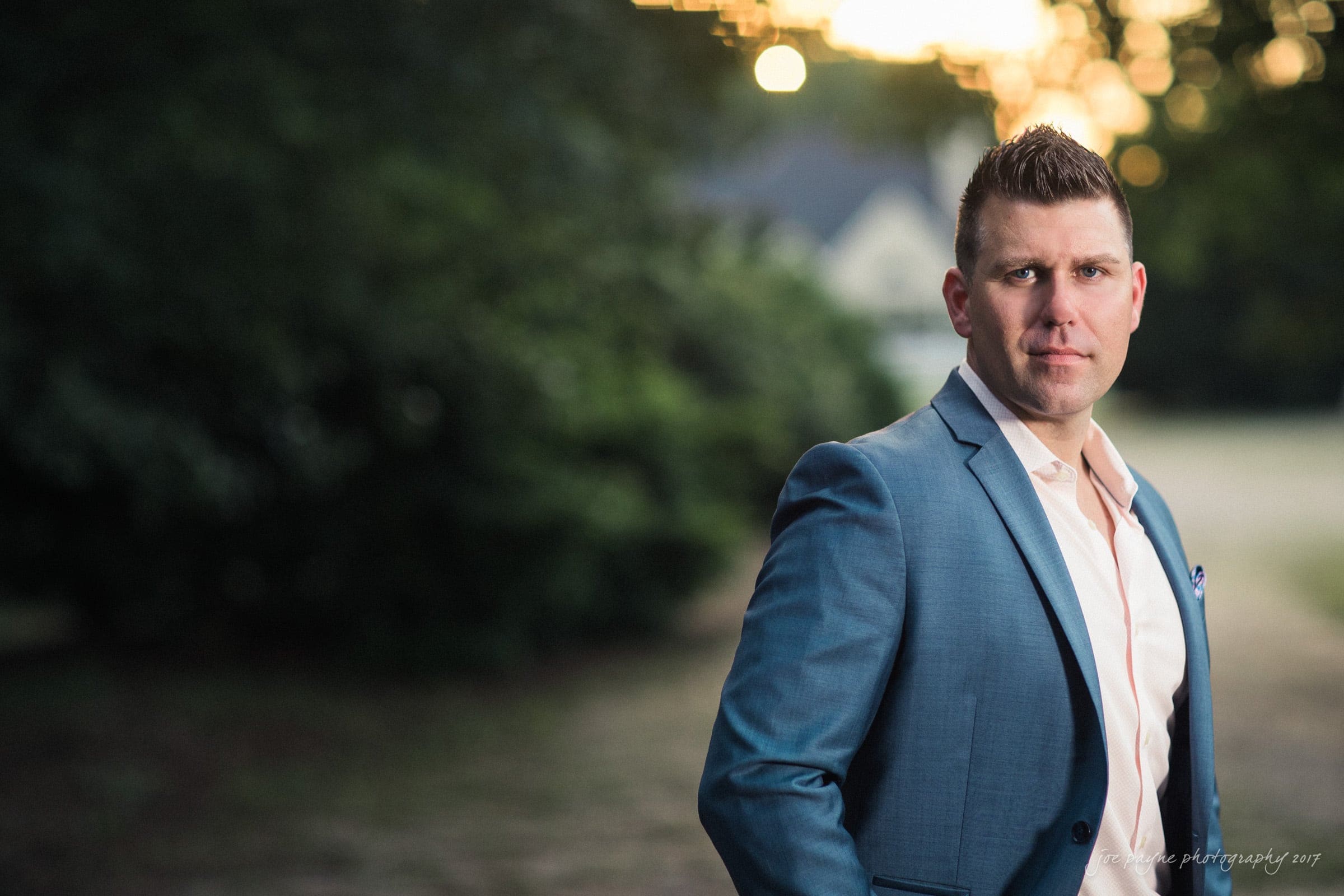 A man in a blue suit stands outdoors with blurred greenery in the background. The lighting suggests its either dusk or dawn, creating a serene atmosphere.