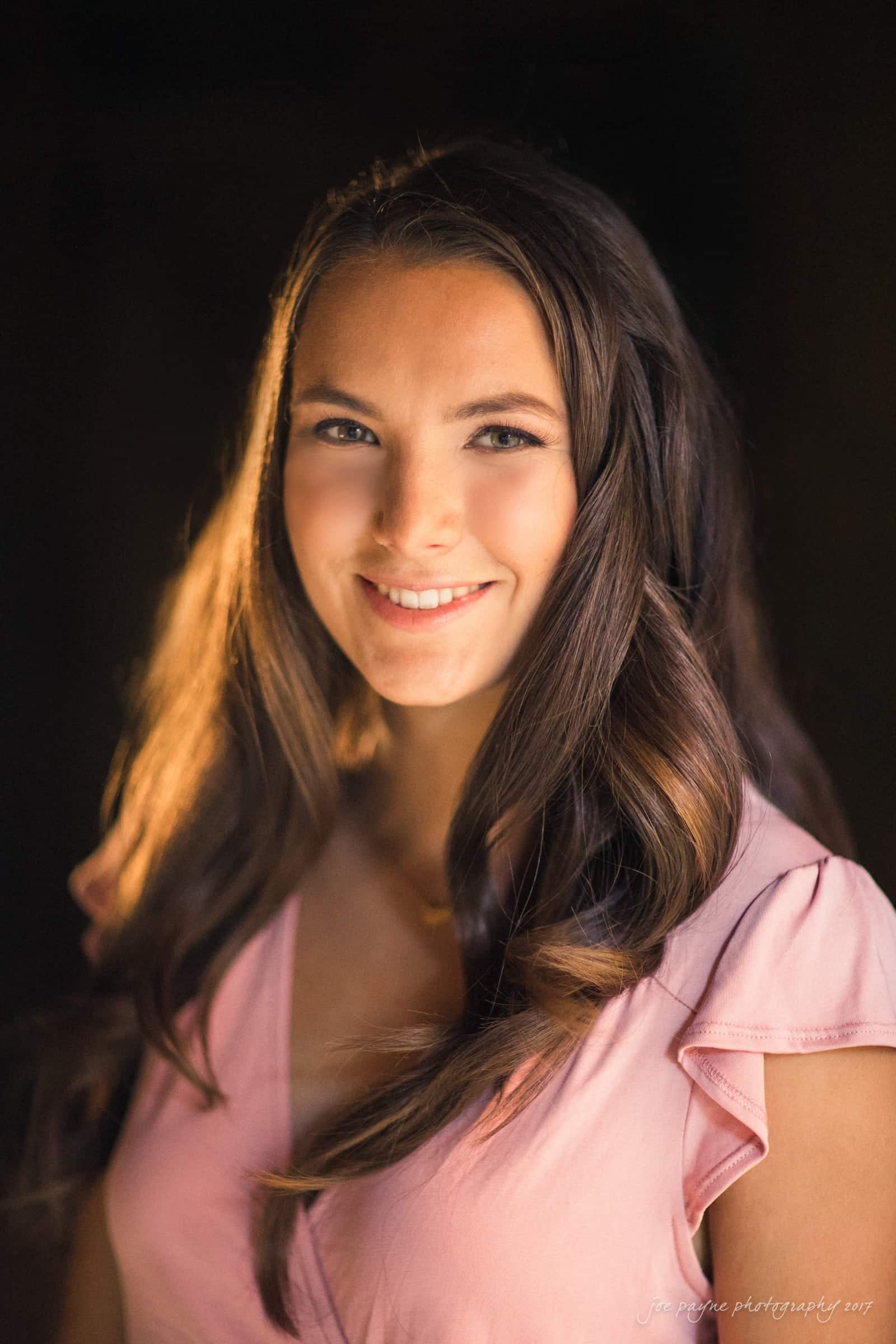 A woman with long, wavy brown hair is smiling softly. She is wearing a light pink blouse and is illuminated by warm, soft lighting, against a dark background.