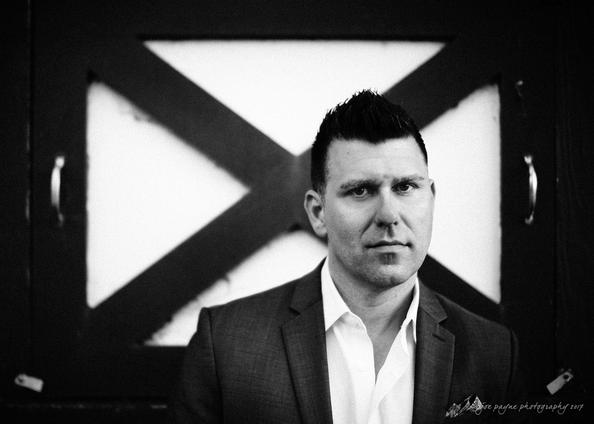 A man in a suit stands in front of a wooden door with an X pattern. The image is in black and white, giving it a classic and focused feel. The background features two metal handles.