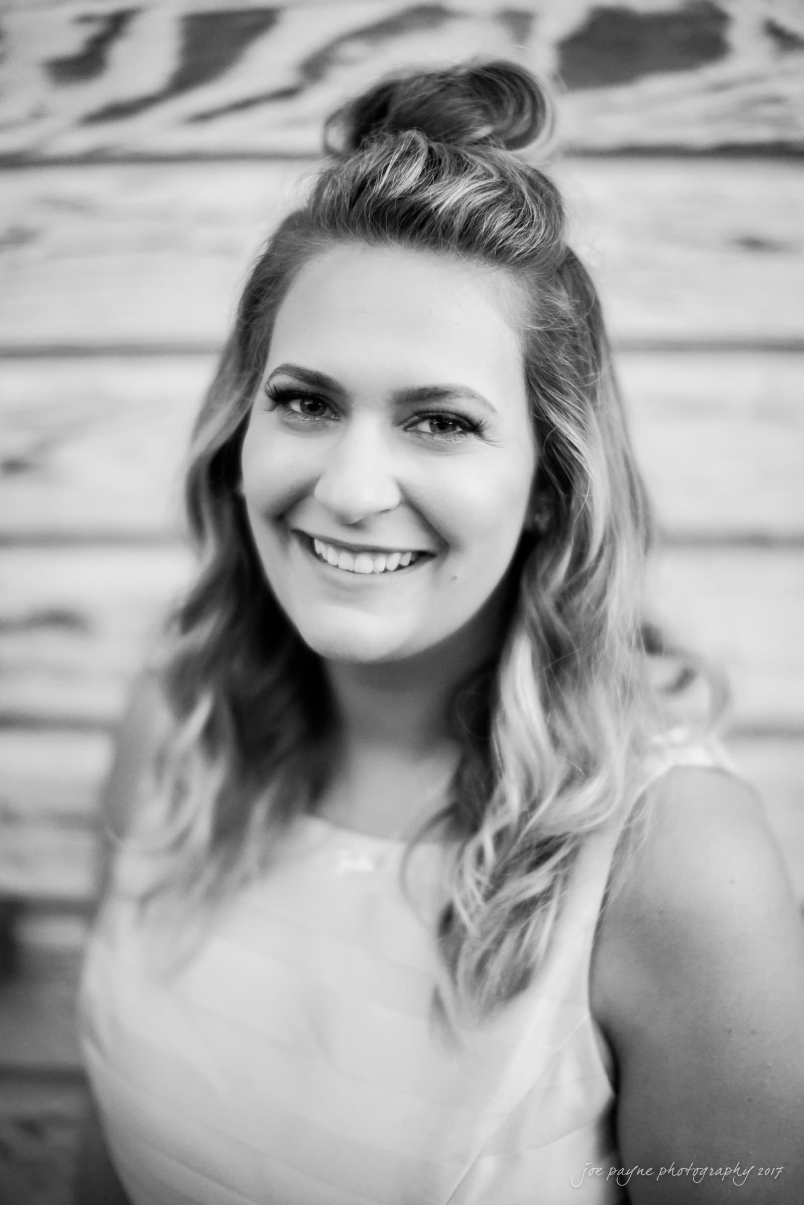 Black and white portrait of a smiling woman with wavy hair styled in a half-up bun, wearing a sleeveless top. She stands in front of a wooden background.