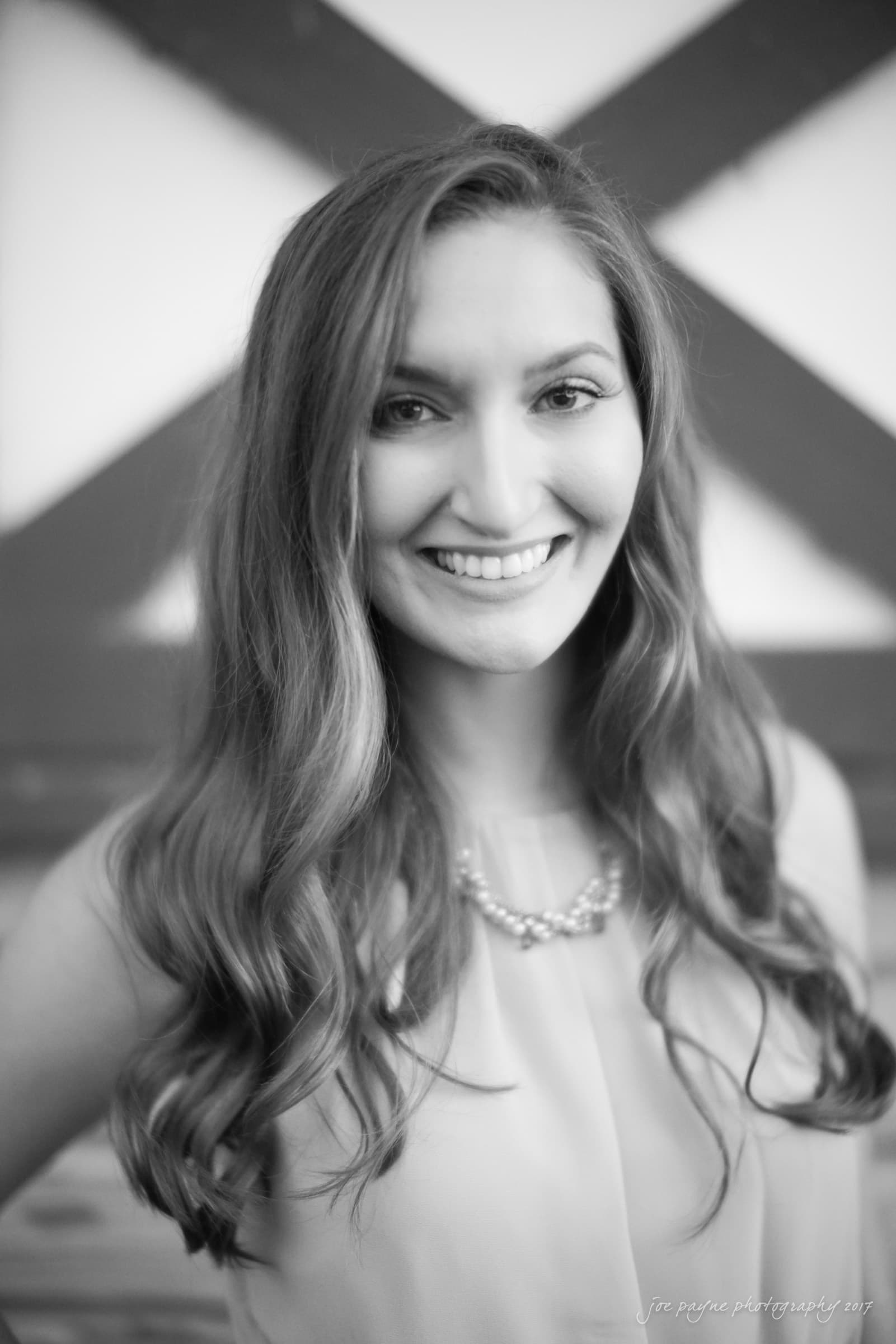 A black and white portrait of a woman smiling. She has long, wavy hair and is wearing a sleeveless top with a necklace. The background features a geometric design.