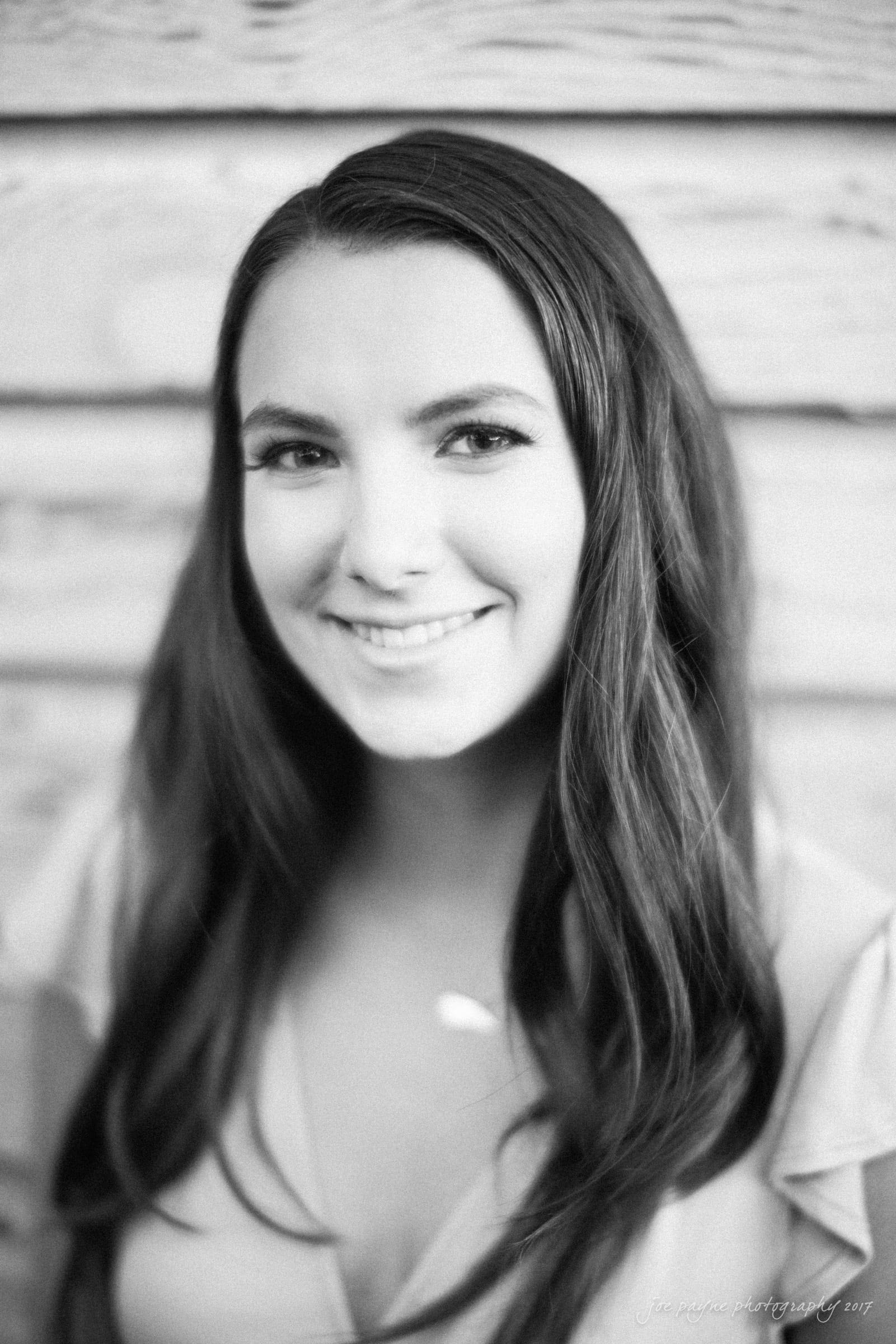 Black and white portrait of a smiling woman with long dark hair, wearing a light-colored top. The background is a wooden texture.