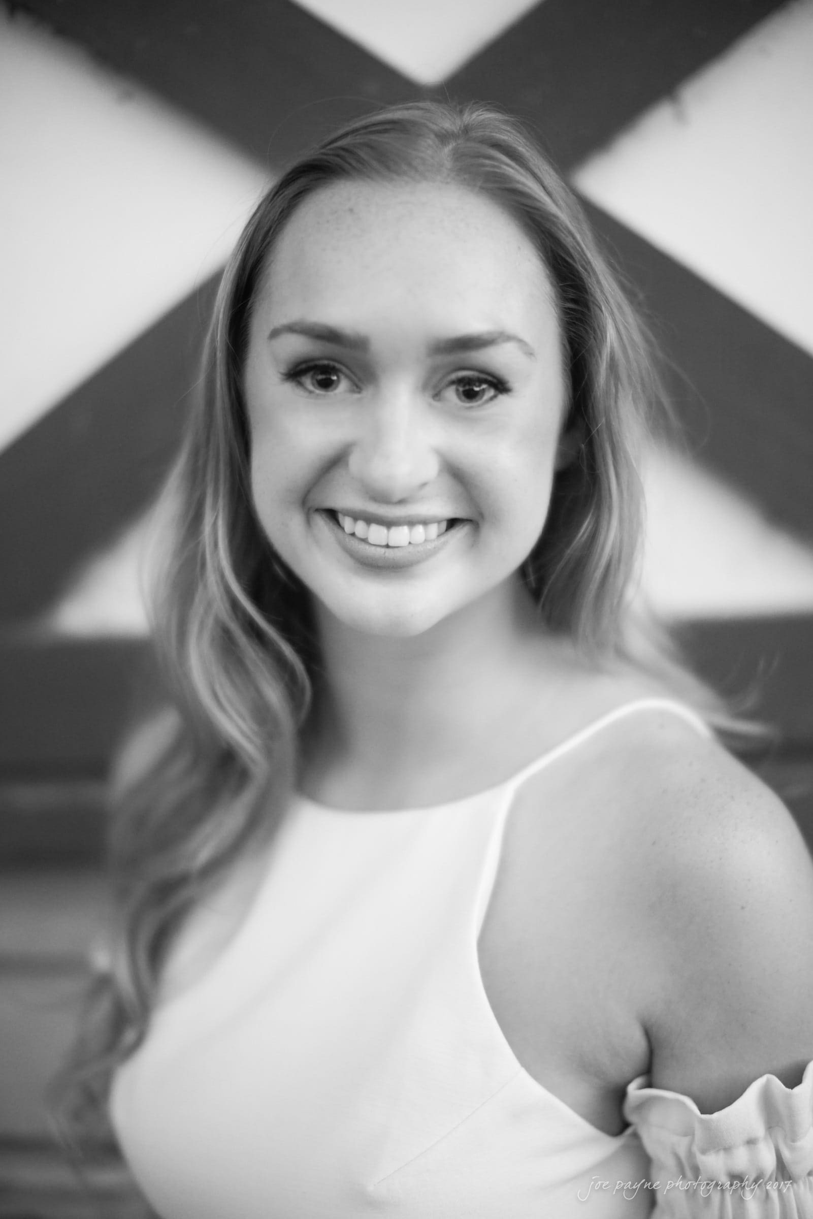Black and white image of a person smiling at the camera, with long hair, wearing a sleeveless top. A blurred geometric pattern appears in the background.