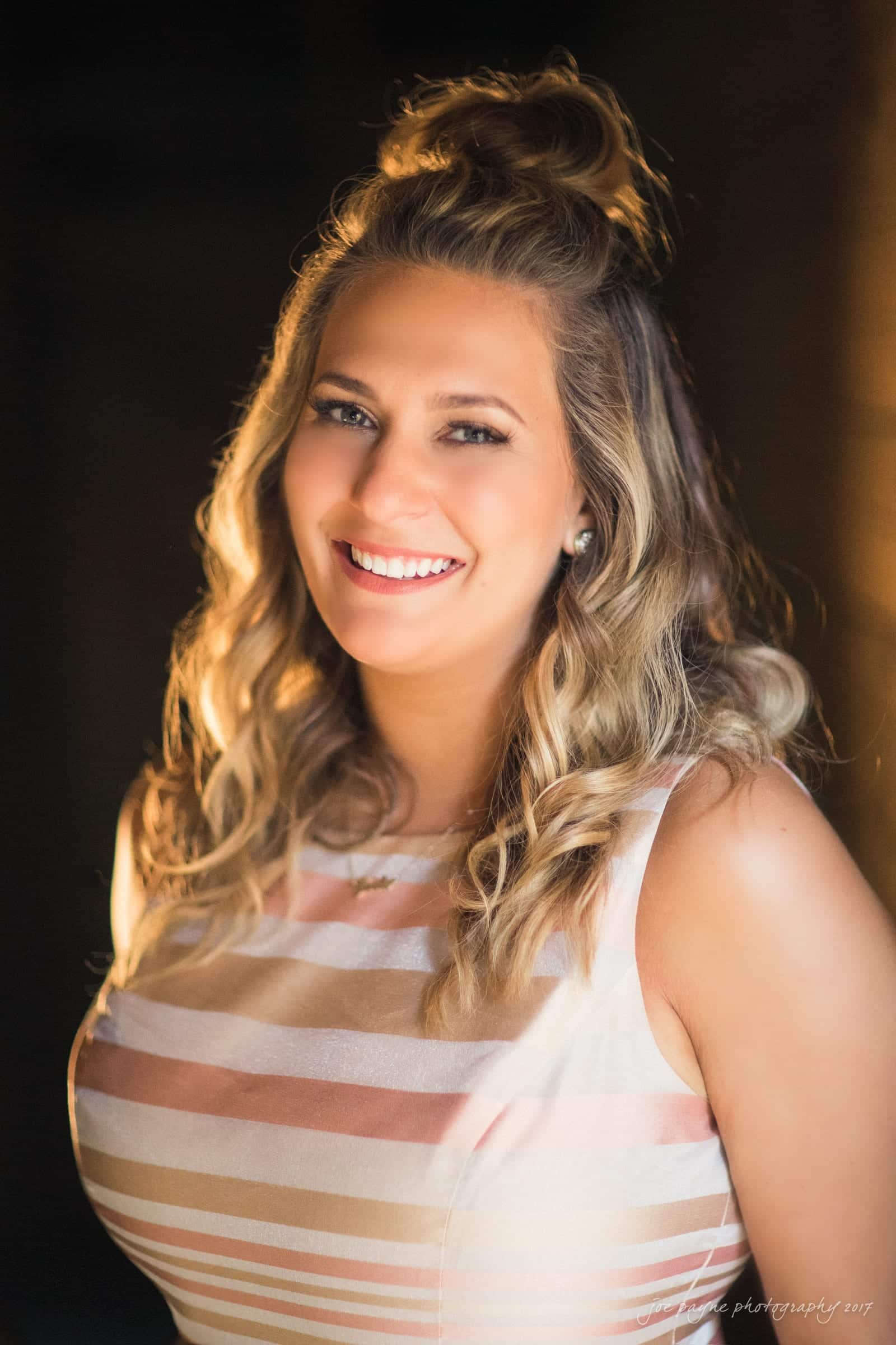 A woman with curly hair smiles warmly. She is wearing a light-colored striped dress and is illuminated by soft lighting. The background is dark, making her stand out.