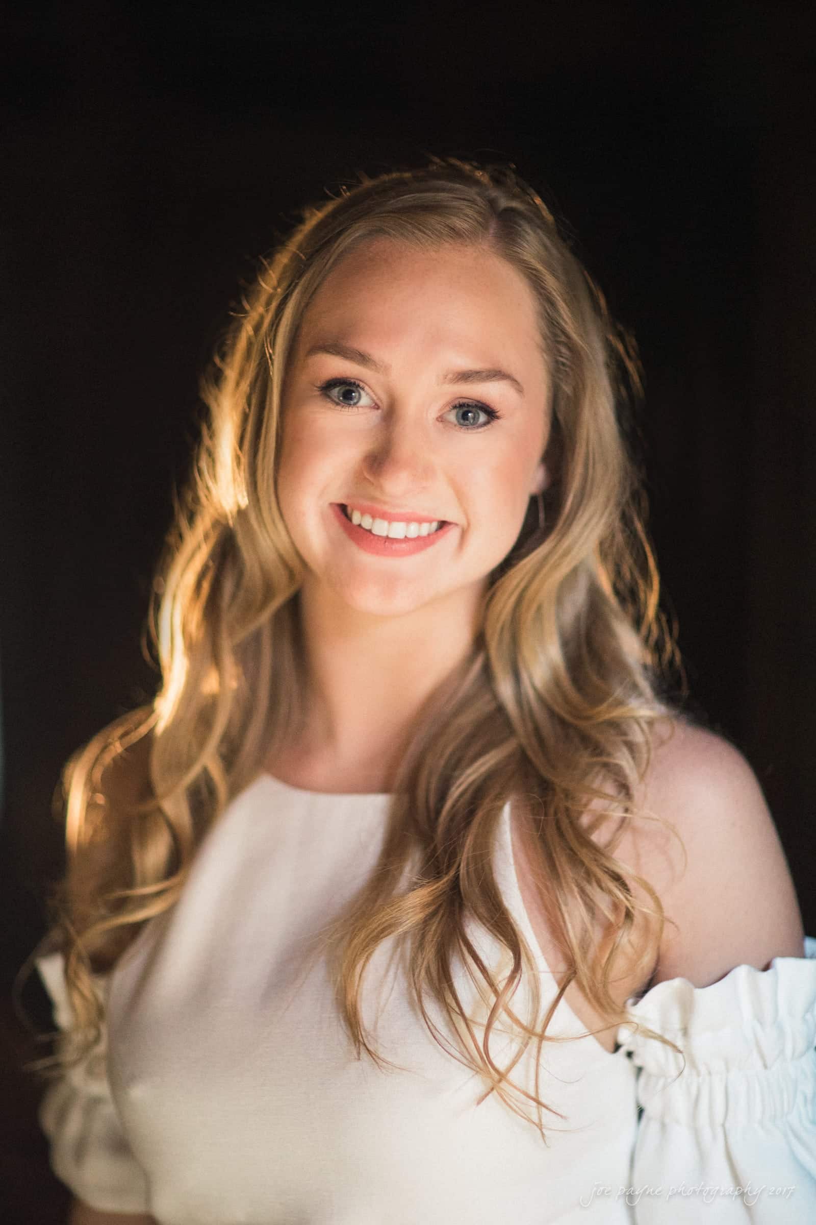 A woman with long blonde hair smiles warmly at the camera. She is wearing a white off-the-shoulder top with soft lighting highlighting her face against a dark background.