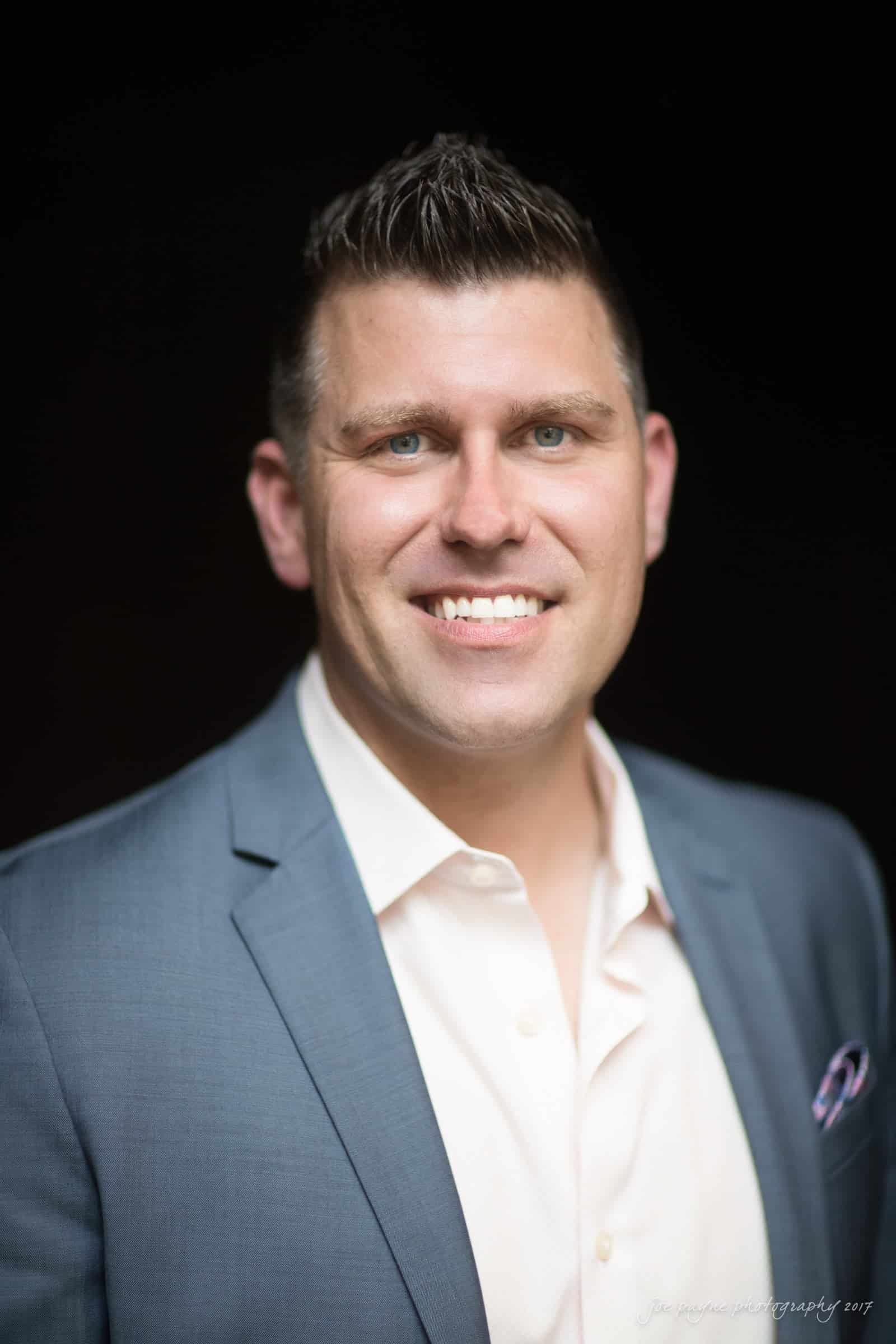 A man with short, dark hair and a bright smile is wearing a light pink shirt and a blue blazer. The background is dark, making him stand out prominently in the image.