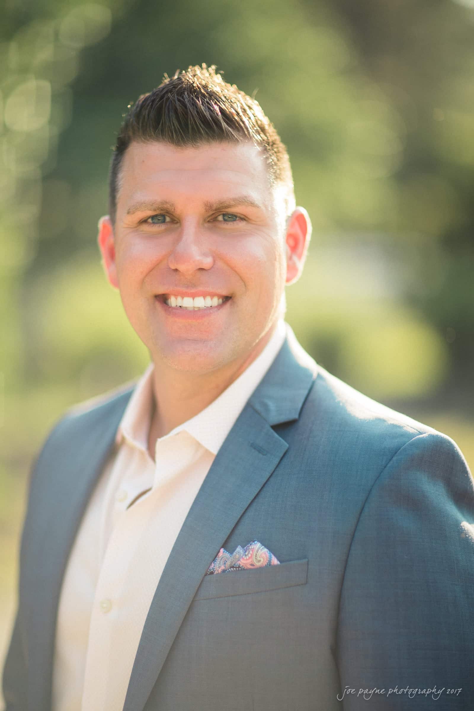 A person in a blue suit jacket and white shirt is smiling brightly. The background is a soft-focus outdoor setting with greenery, and the lighting creates a warm, sunny atmosphere.