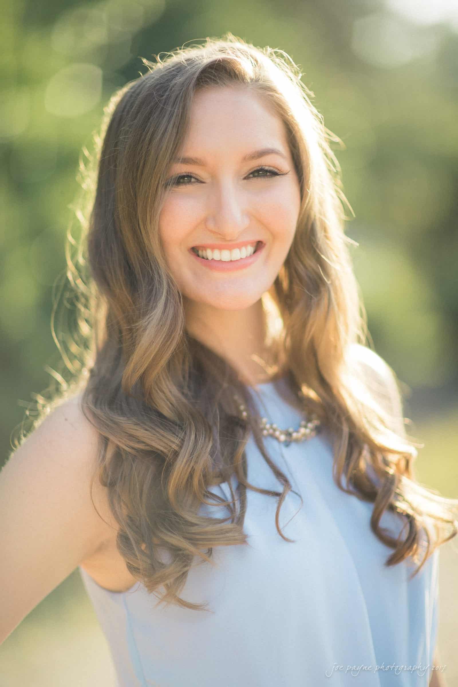 A woman with long, wavy hair smiles brightly while standing outdoors. She is wearing a light sleeveless top and a necklace. The background is softly blurred with greenery and sunlight creating a warm, glowing effect.