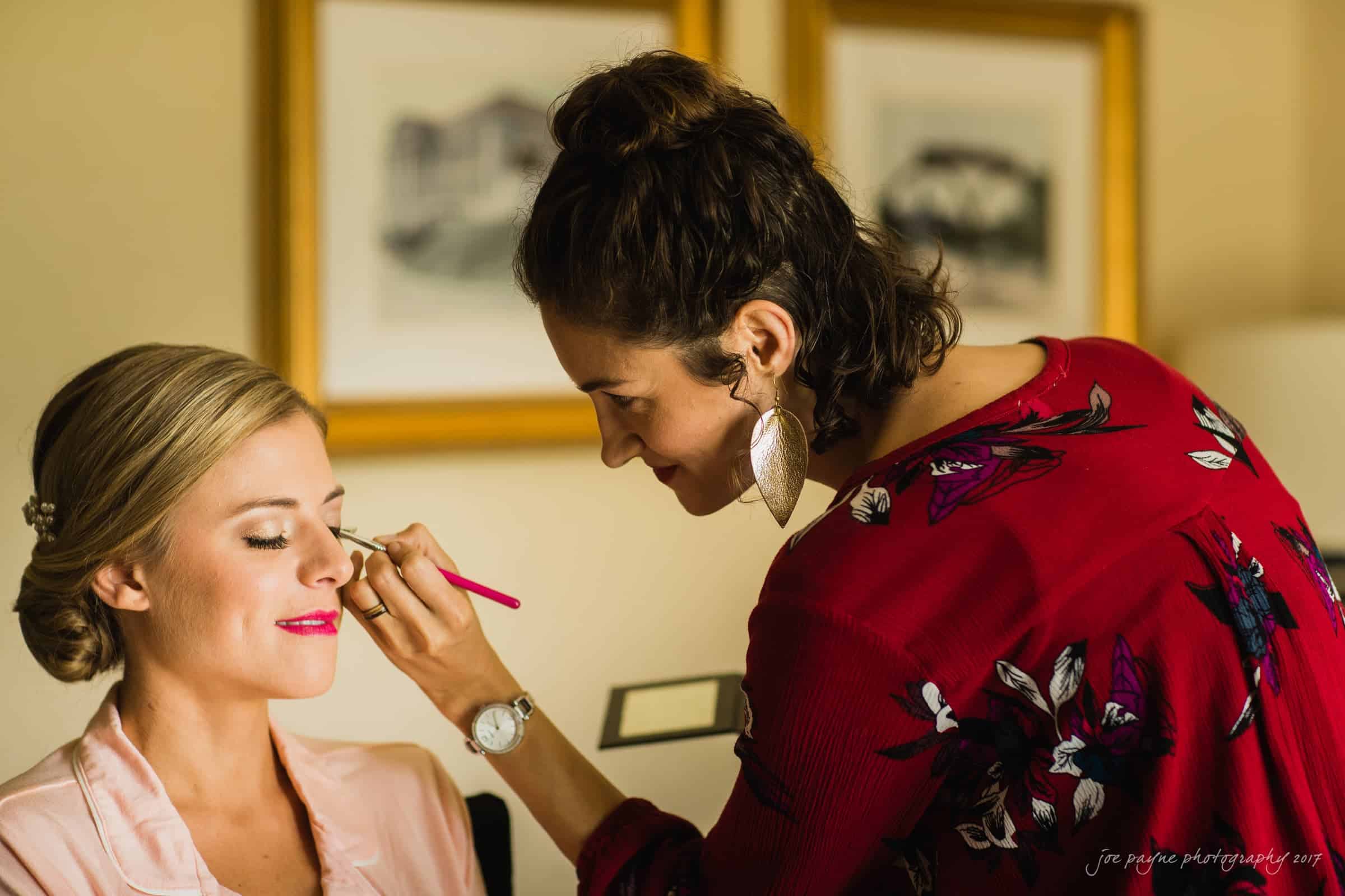 A makeup artist is applying eye makeup to a smiling woman with blonde hair in an updo. The artist is wearing a red, floral-patterned top and large earrings. The background features framed artwork on the wall.