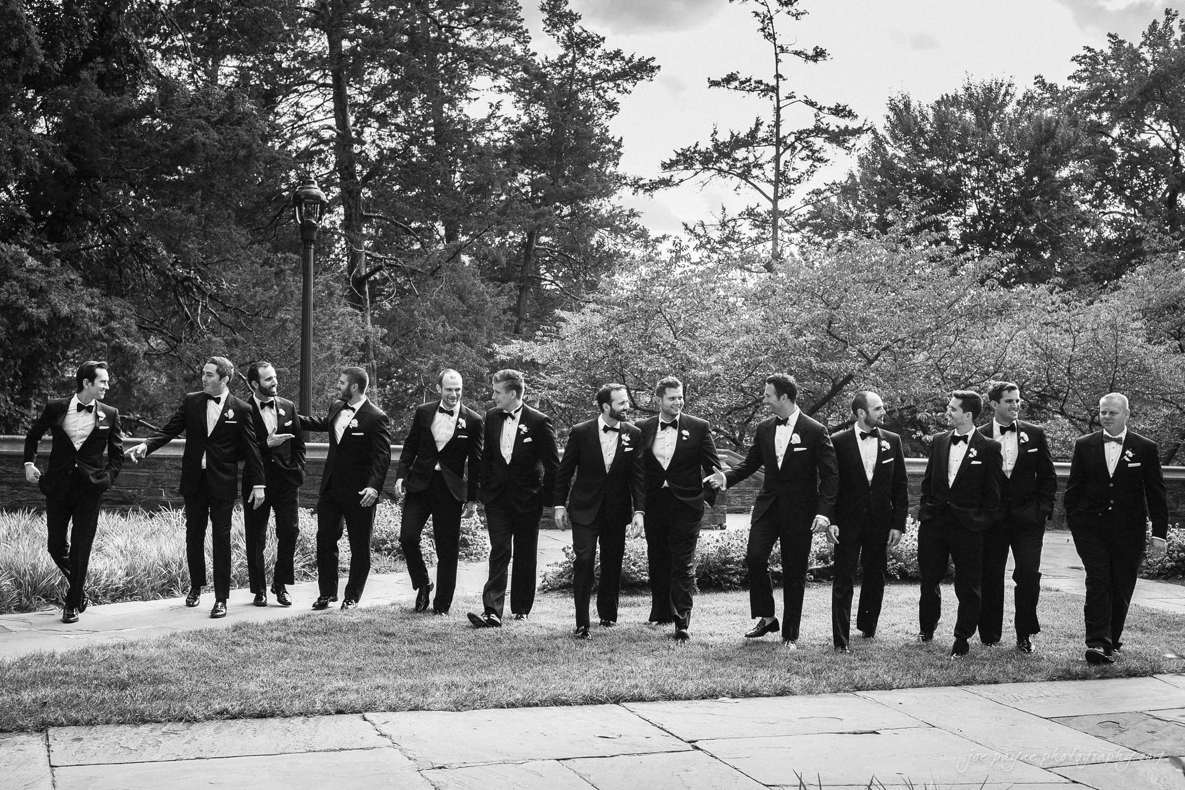 A black and white photo of thirteen men in tuxedos walking in a line on a stone pathway. They are outdoors, with trees and grass in the background. Some are smiling and holding hands, creating a joyful and relaxed atmosphere.