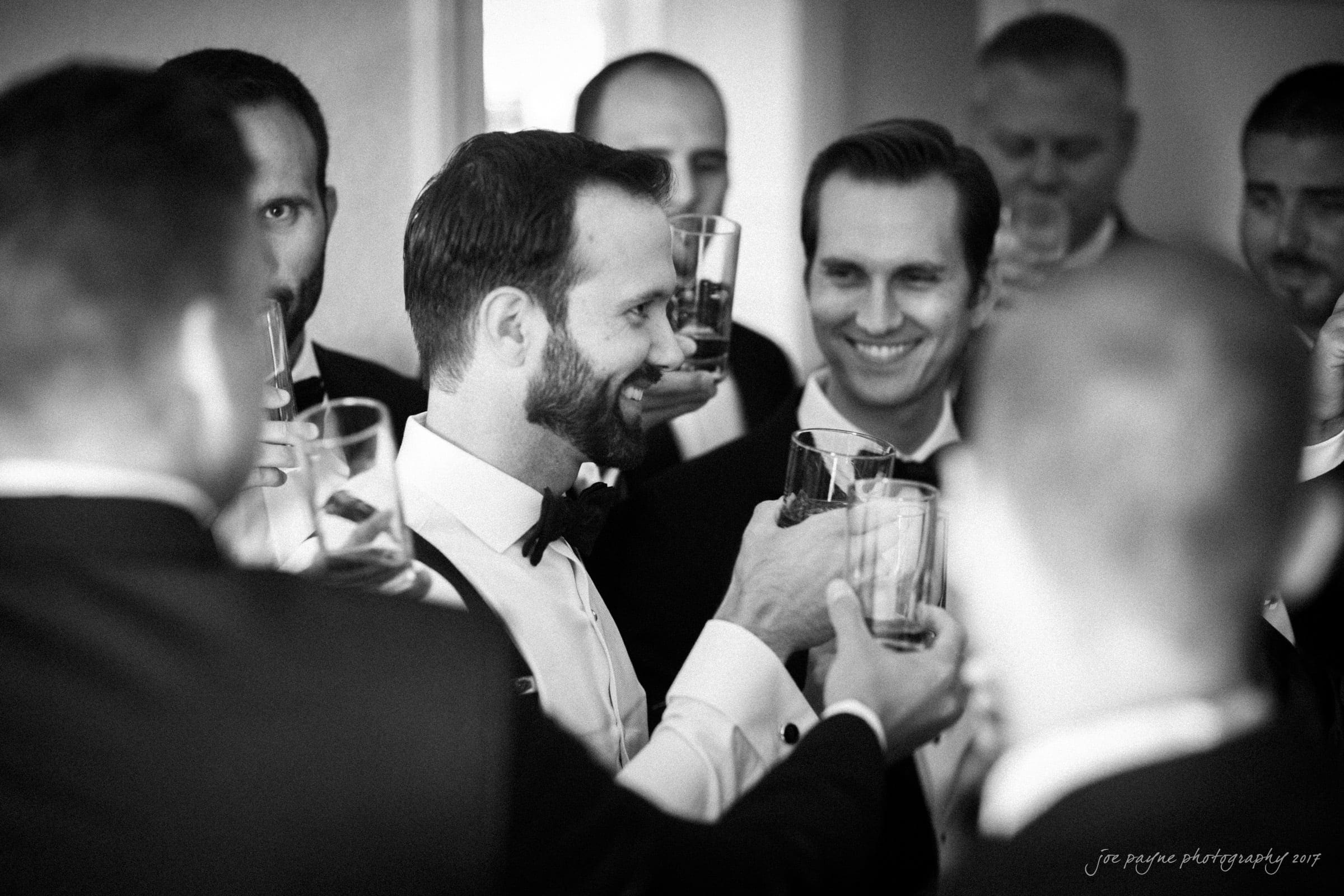 A group of men in tuxedos are clinking glasses in celebration, smiling and standing closely together in a well-lit room. The mood is festive and joyful.