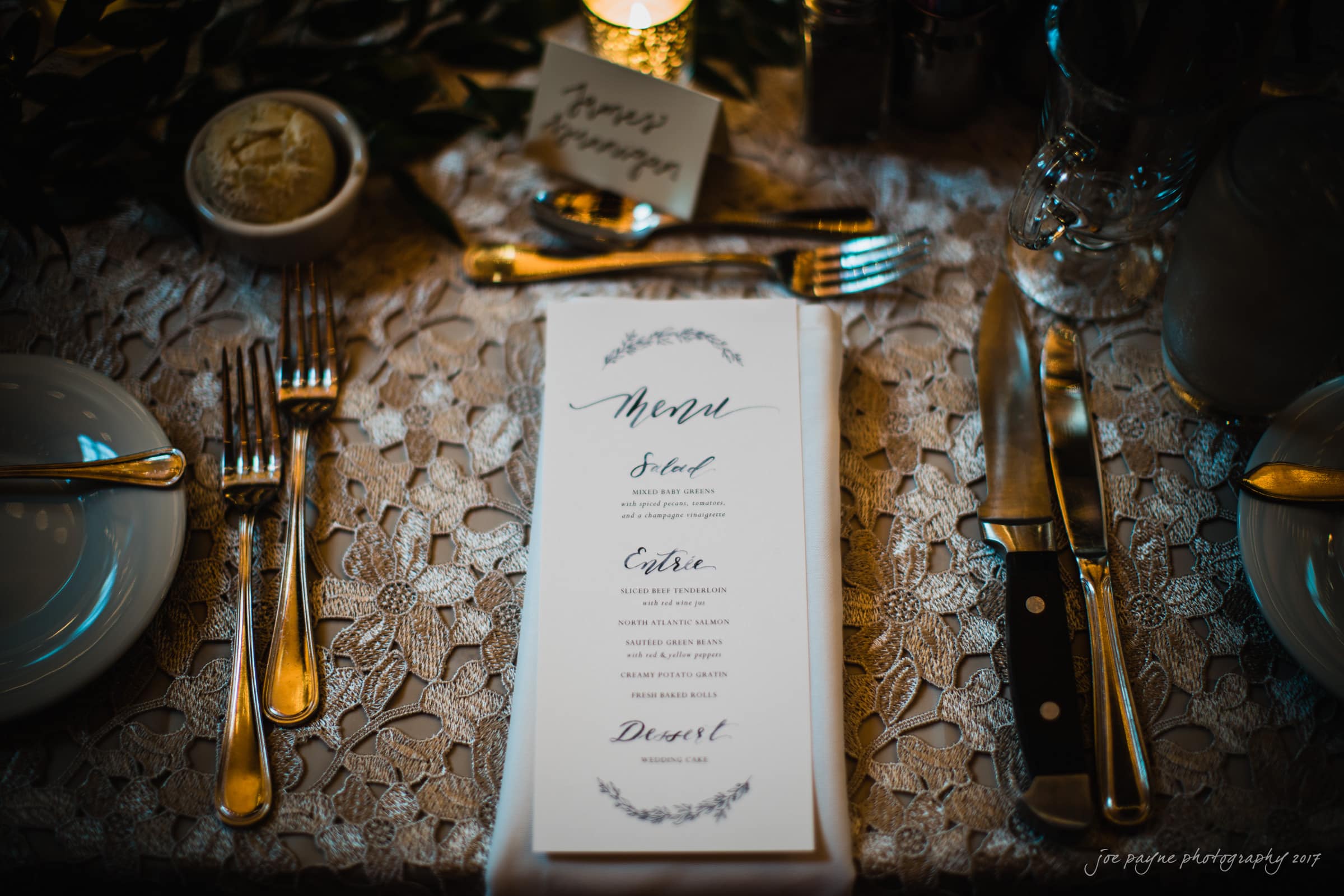 A rustic table setting with a detailed lace tablecloth, elegant menu, polished silverware, and a small bowl of butter. A candle flickers nearby, casting a warm glow over the sophisticated arrangement.