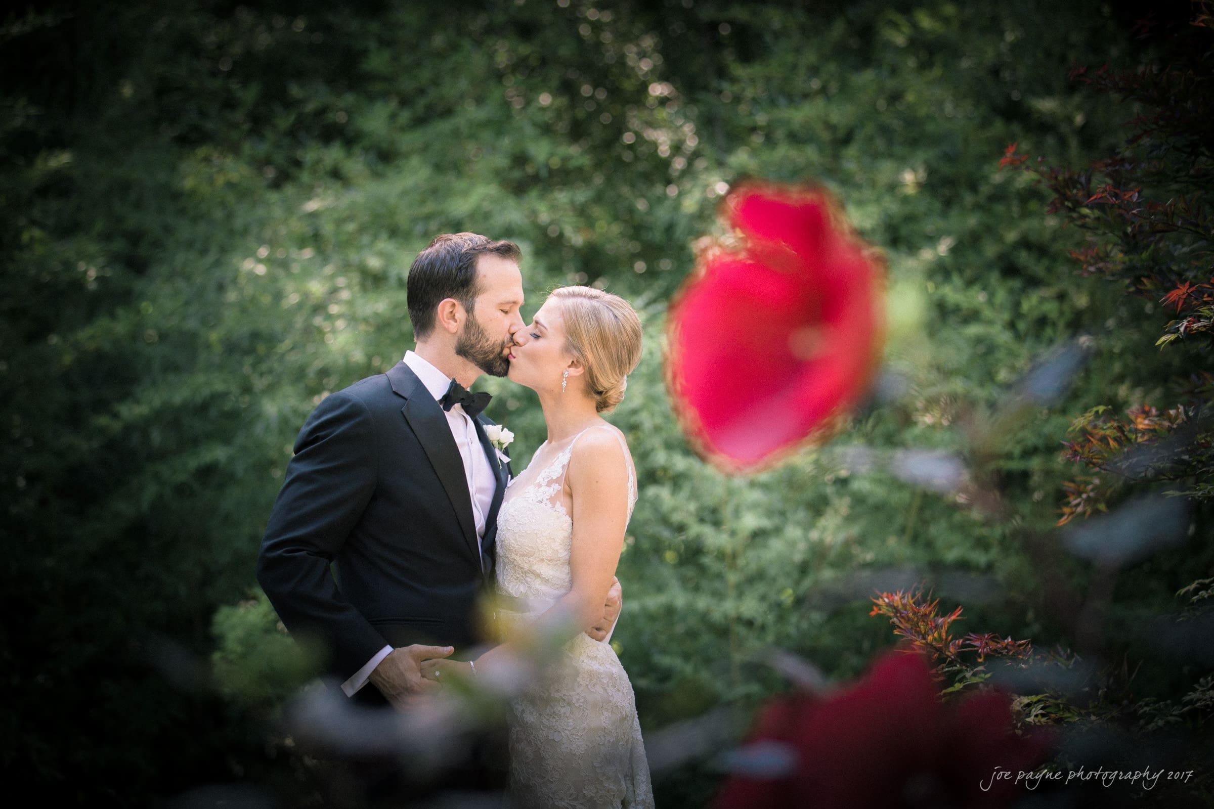 A couple in wedding attire shares a kiss in a garden setting, surrounded by lush greenery. A red flower is prominently blurred in the foreground.