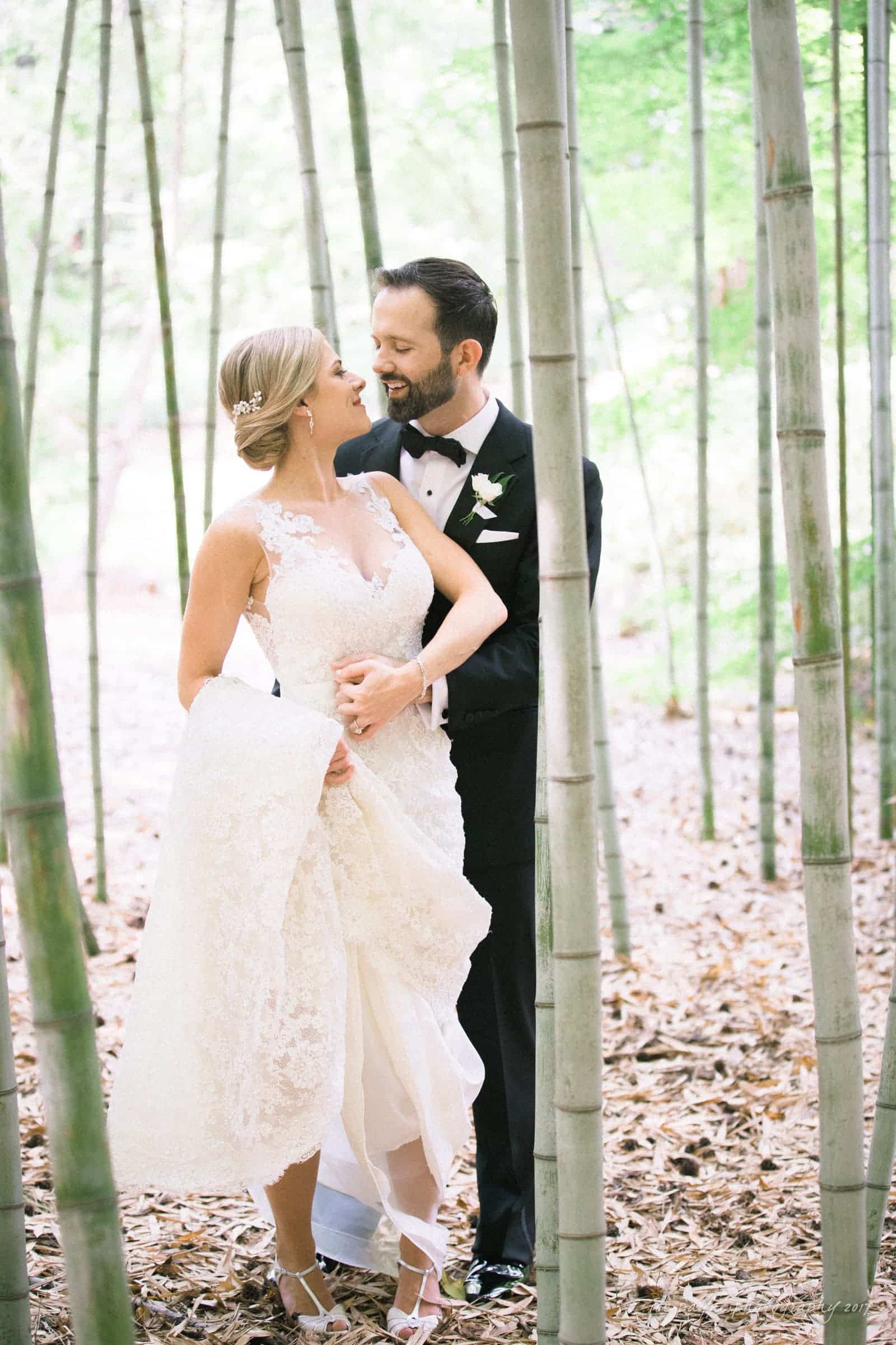 A couple in wedding attire stands together in a serene bamboo forest. The bride, in a lace gown, holds her dress while looking at the groom, who wears a black tuxedo. They are smiling and surrounded by tall bamboo stalks.