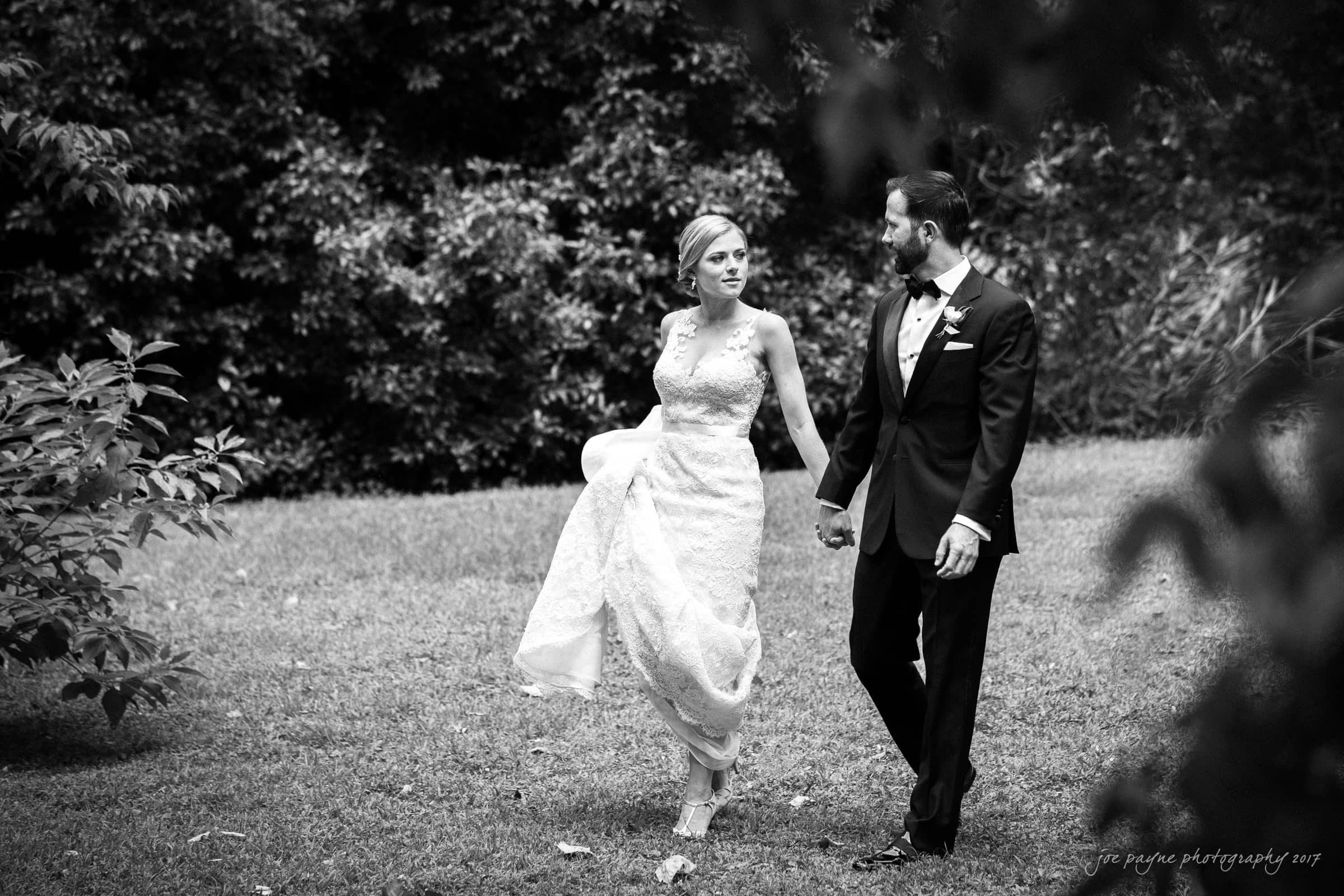 A black and white photo of a bride and groom holding hands while walking on grass. The bride wears a long lace dress, and the groom is in a suit with a bow tie. Trees and bushes are in the background.