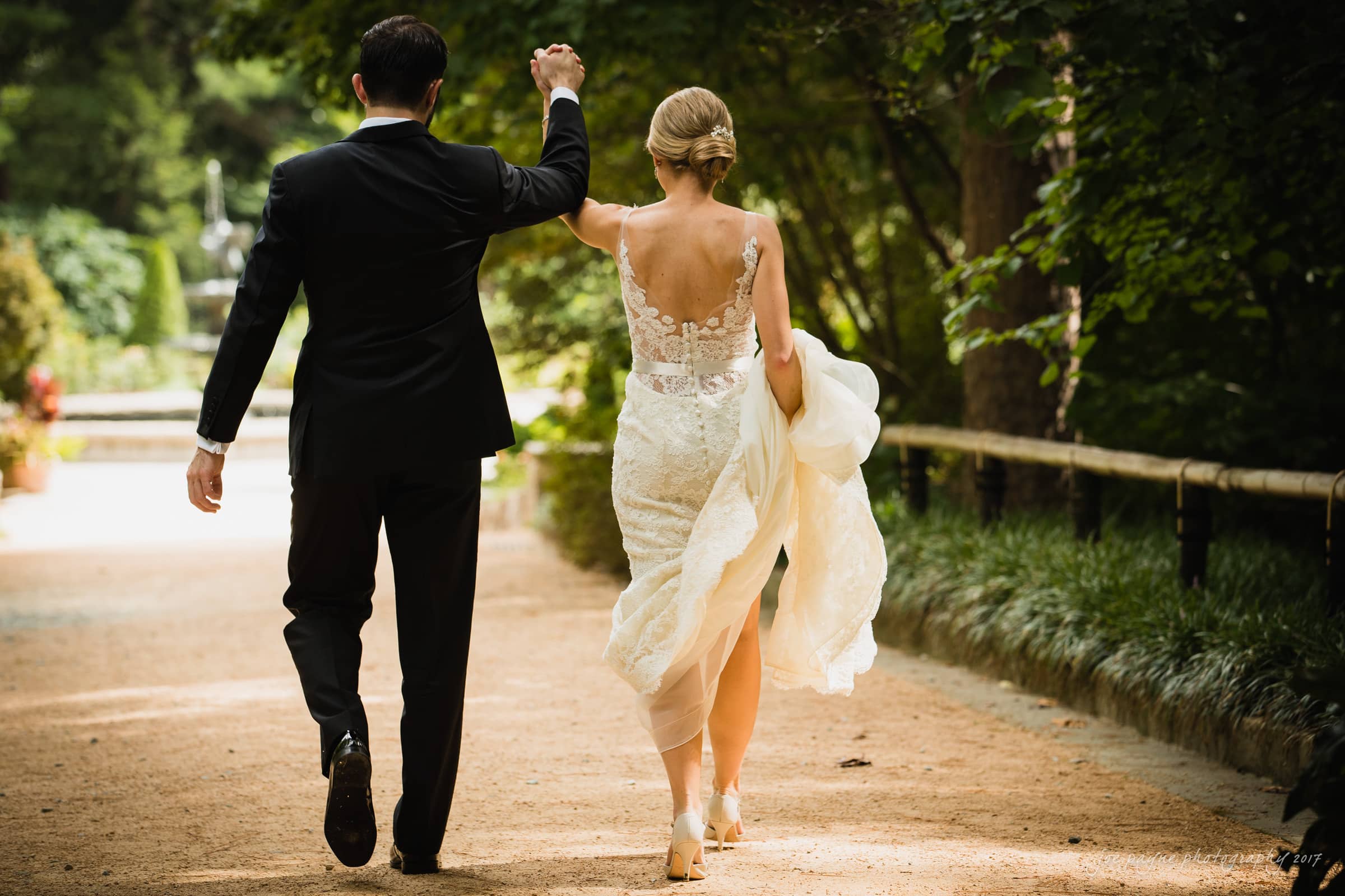 A couple walks hand in hand down a tree-lined path. The woman wears a white lace gown, and the man is in a dark suit. Their backs are turned to the camera, and the setting is lush and green, suggesting a garden or park.