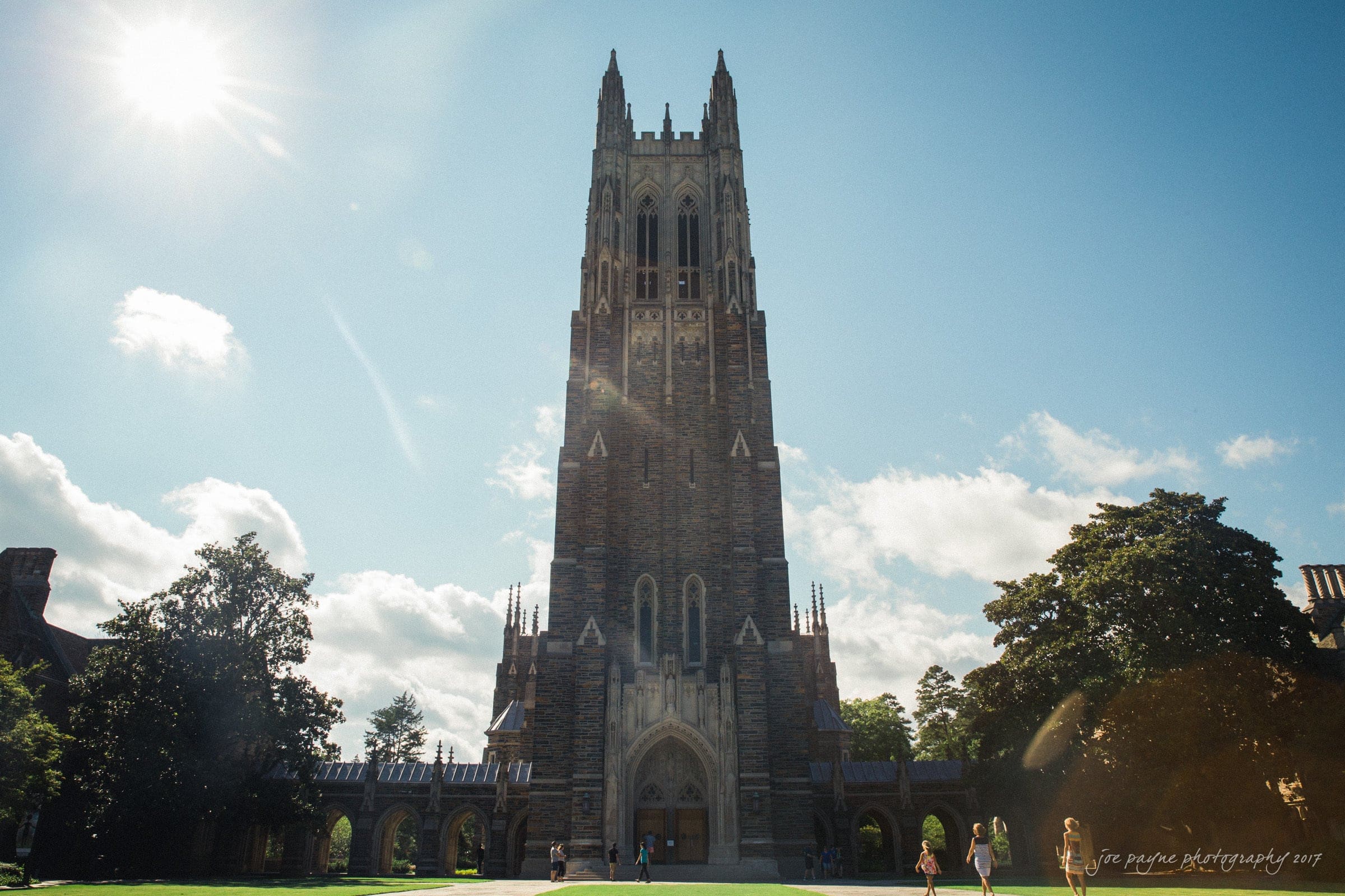 A tall, Gothic-style chapel stands against a clear blue sky, surrounded by green trees and grass. People walk around the sunny courtyard in front of the chapel.