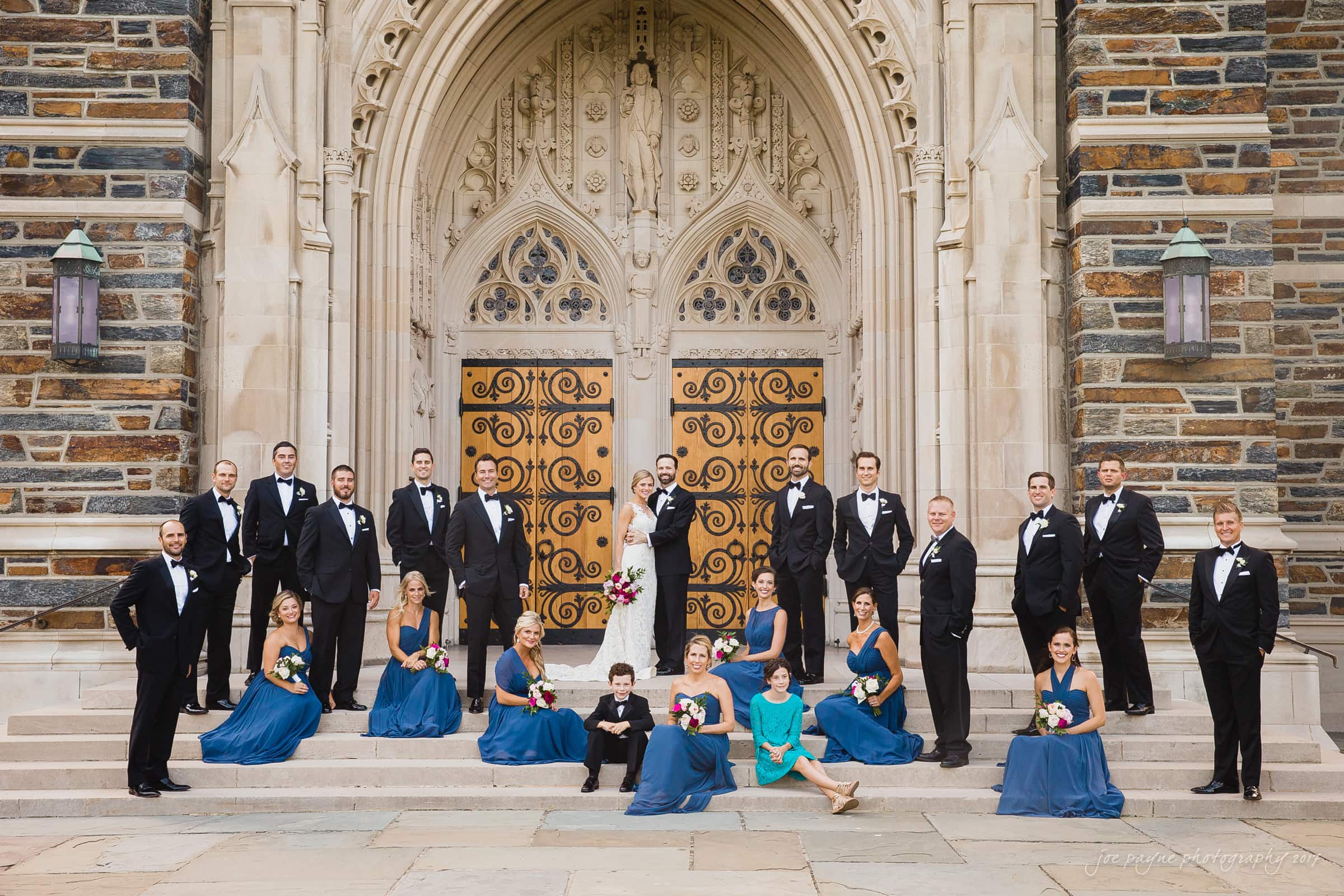 Duke Chapel Weddings A wide-angle bridal party group portrait on the stone steps of Duke Chapel, with the bridesmaids in blue dresses seated, the groomsmen standing, and the smiling bride and groom in the center framed by the ornate church doors.