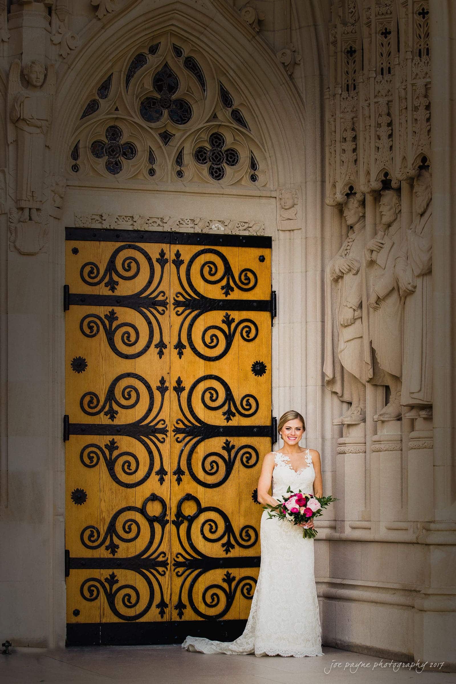 A bride in a lace wedding dress holds a bouquet while standing beside ornate wooden doors with black scrollwork. The stone archway and carved figures in the background create an elegant setting.