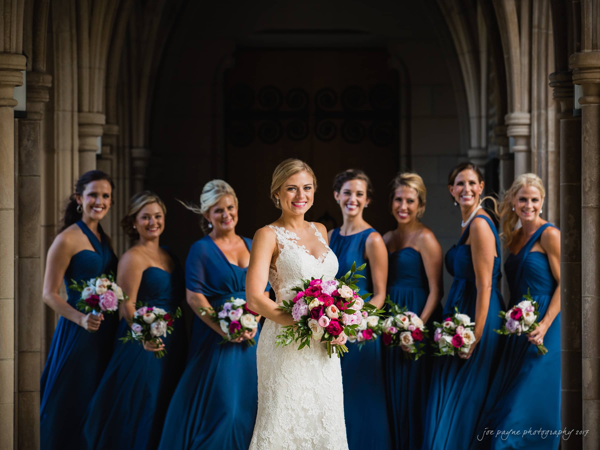 A bride in a white gown stands smiling, holding a bouquet of pink and white flowers. Behind her, six bridesmaids wearing blue dresses also hold similar bouquets. They are posing in an elegant, arched hallway.