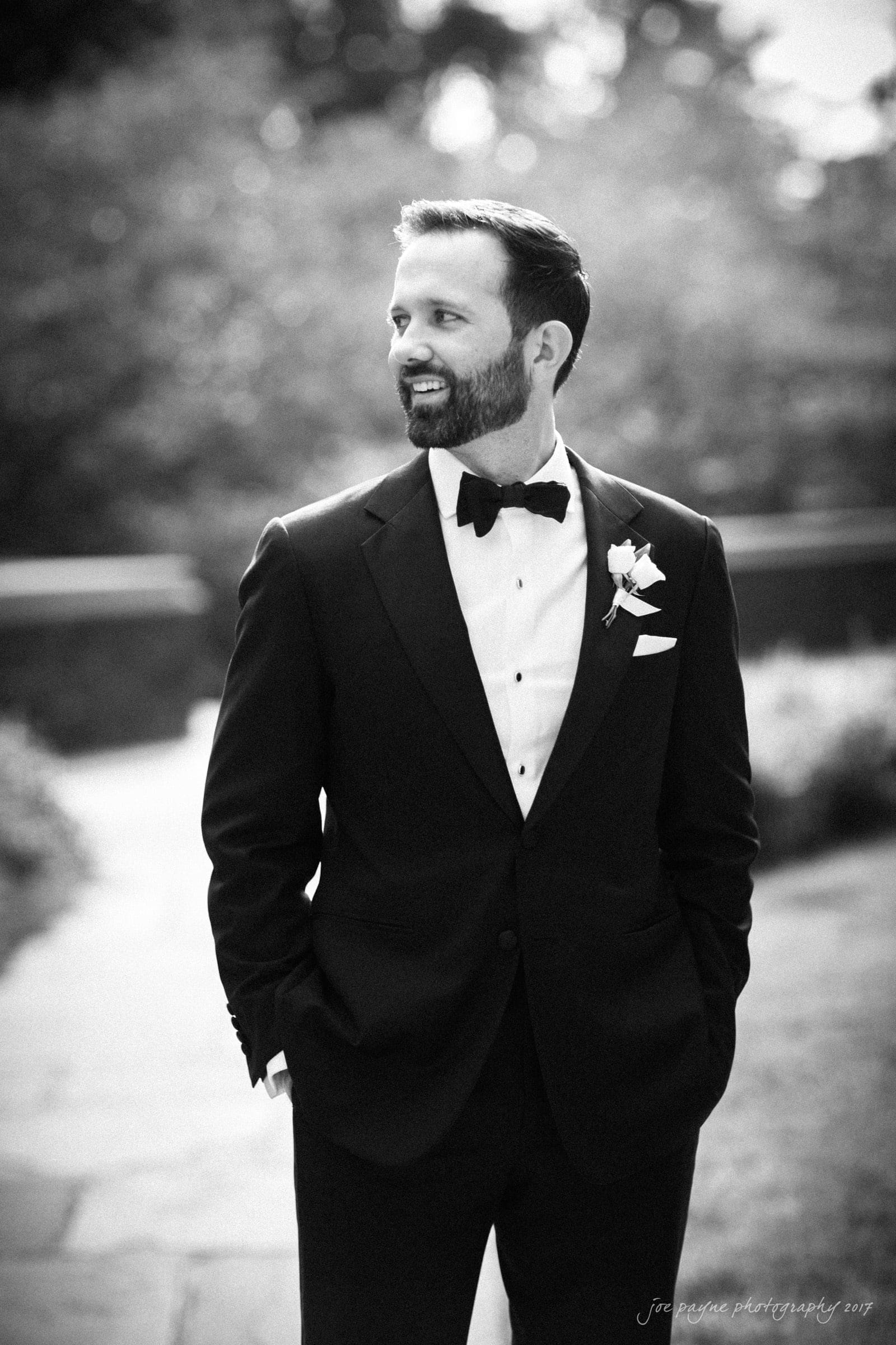 A groom in a tuxedo, complete with a bow tie and boutonniere, stands outside on a stone path. His hands are in his pockets, and he is looking to the side with a smile. The background is softly blurred, creating a classic, elegant atmosphere.