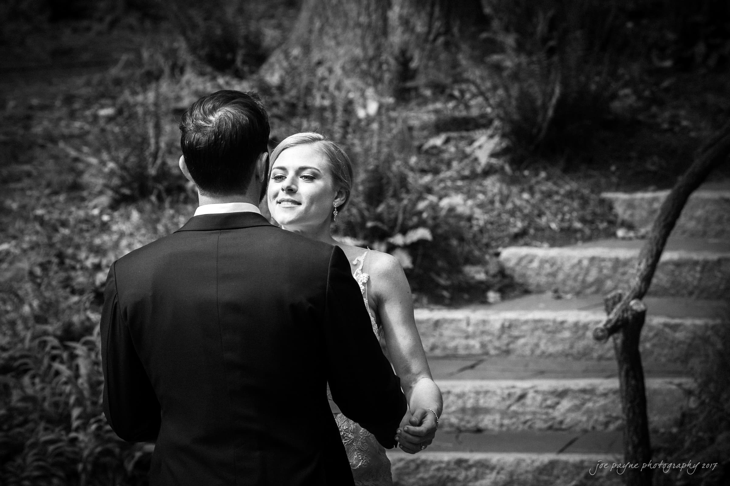 A black and white photo of a couple dancing outdoors on stone steps. The woman smiles at the man, who wears a suit. Trees and plants surround them, creating a natural backdrop.