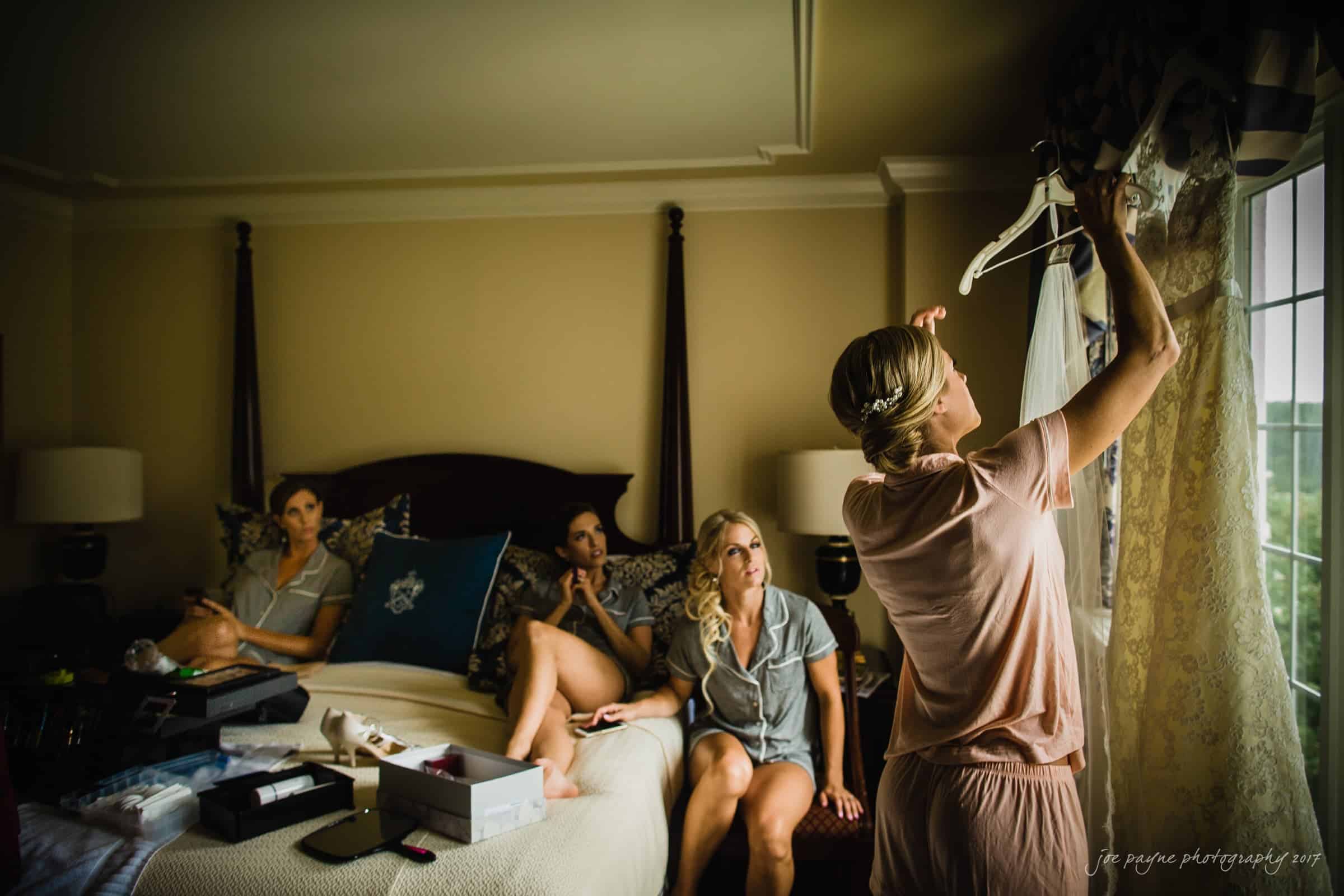 A woman in pajamas examines a wedding dress by a window. Three more women in loungewear sit on a bed watching, with various items around them. The room is softly lit and cozy, suggesting a relaxed wedding preparation scene.