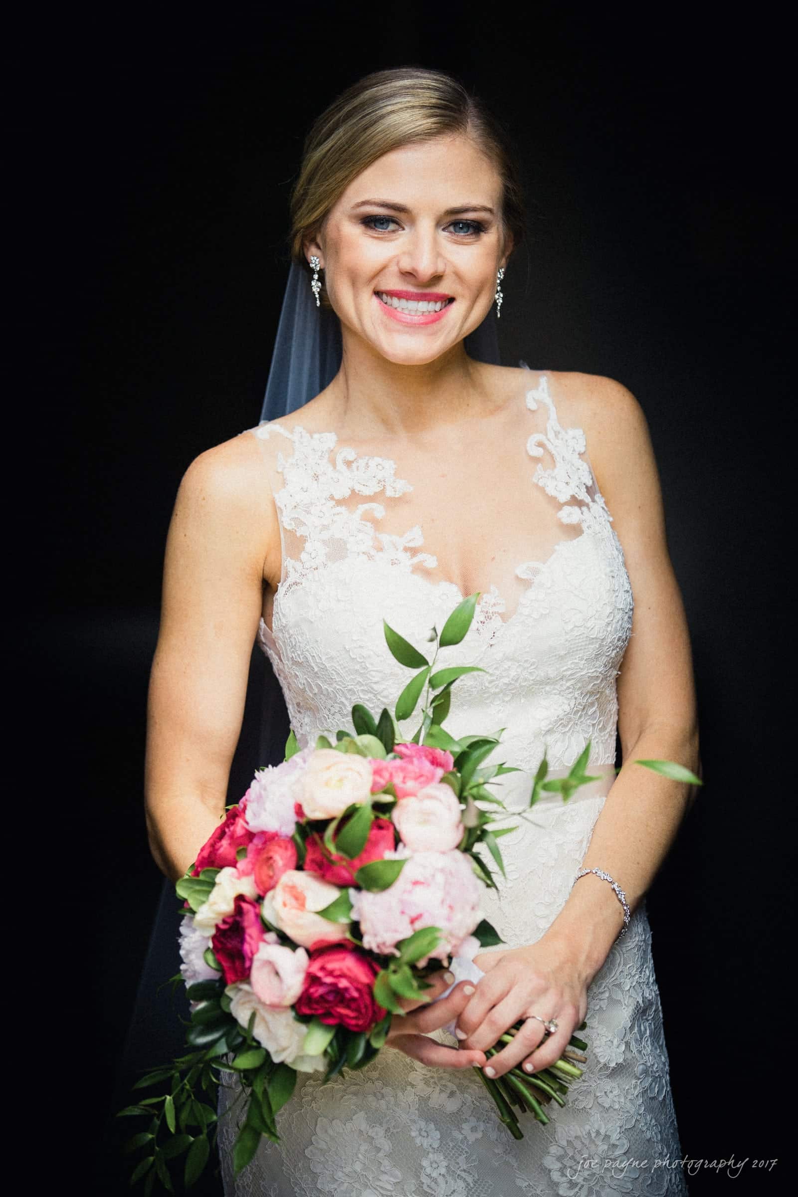 A smiling bride in a white lace wedding dress holds a bouquet of pink and white flowers with greenery. She has her hair styled up and is wearing delicate earrings against a dark background.