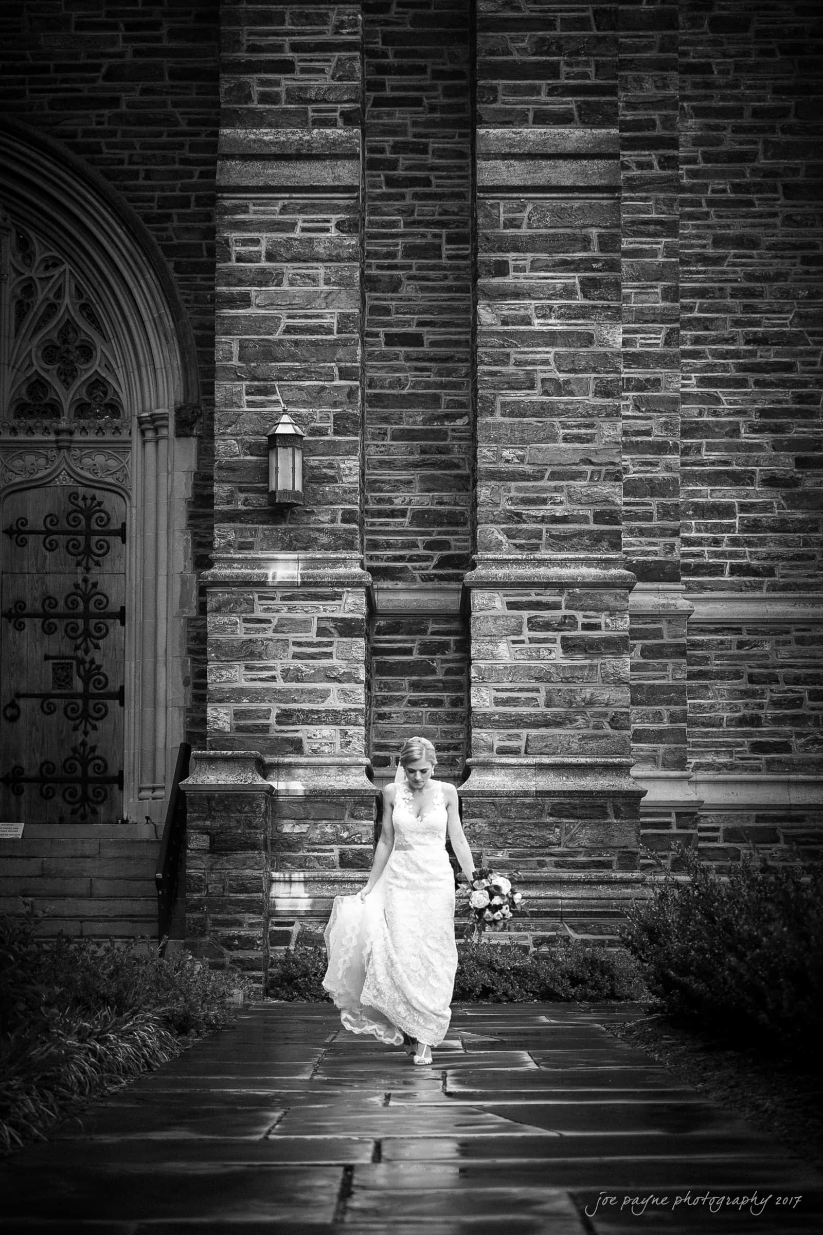 A bride in a white dress walks gracefully on a stone path outside a large, textured stone building with ornate doors. She holds a bouquet in her left hand and lifts her dress slightly with her right hand, smiling softly.