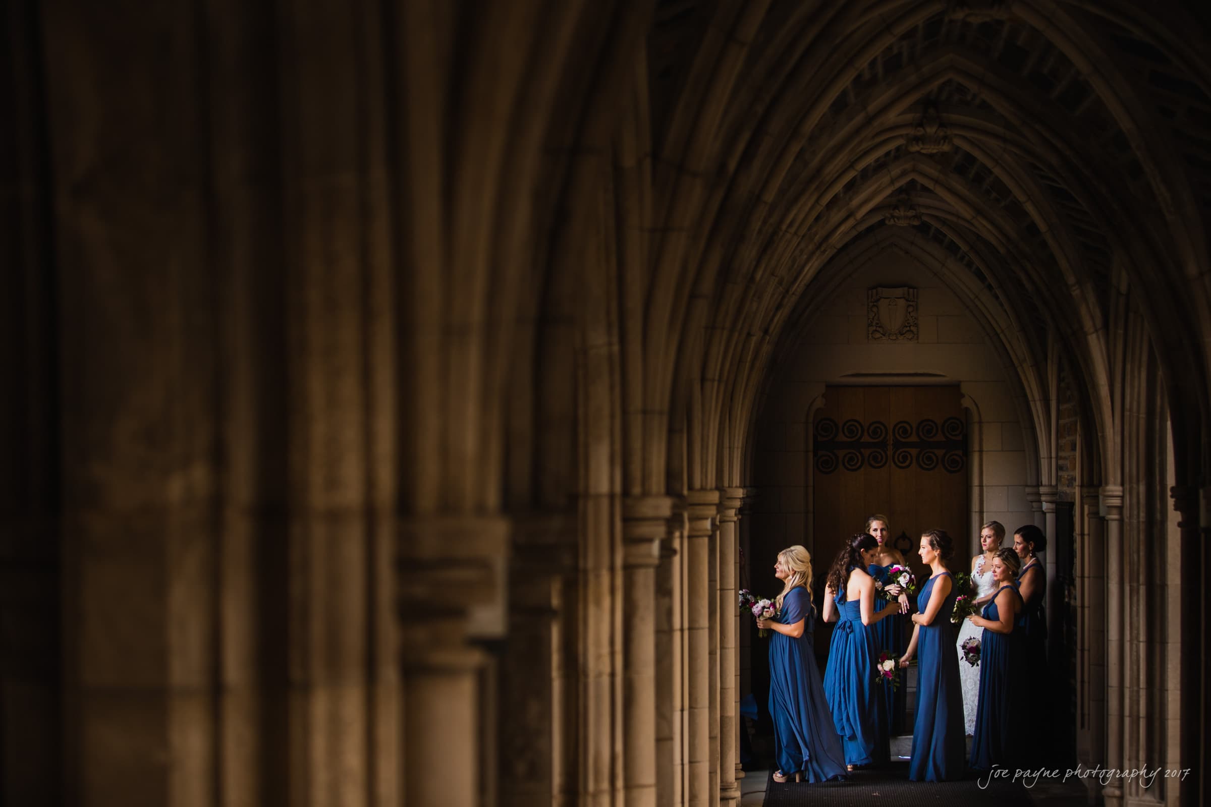 A group of bridesmaids in blue dresses stands in a dimly lit, arched stone hallway. They hold bouquets and appear to be waiting or observing something out of frame. The architectural details create a dramatic, elegant atmosphere.