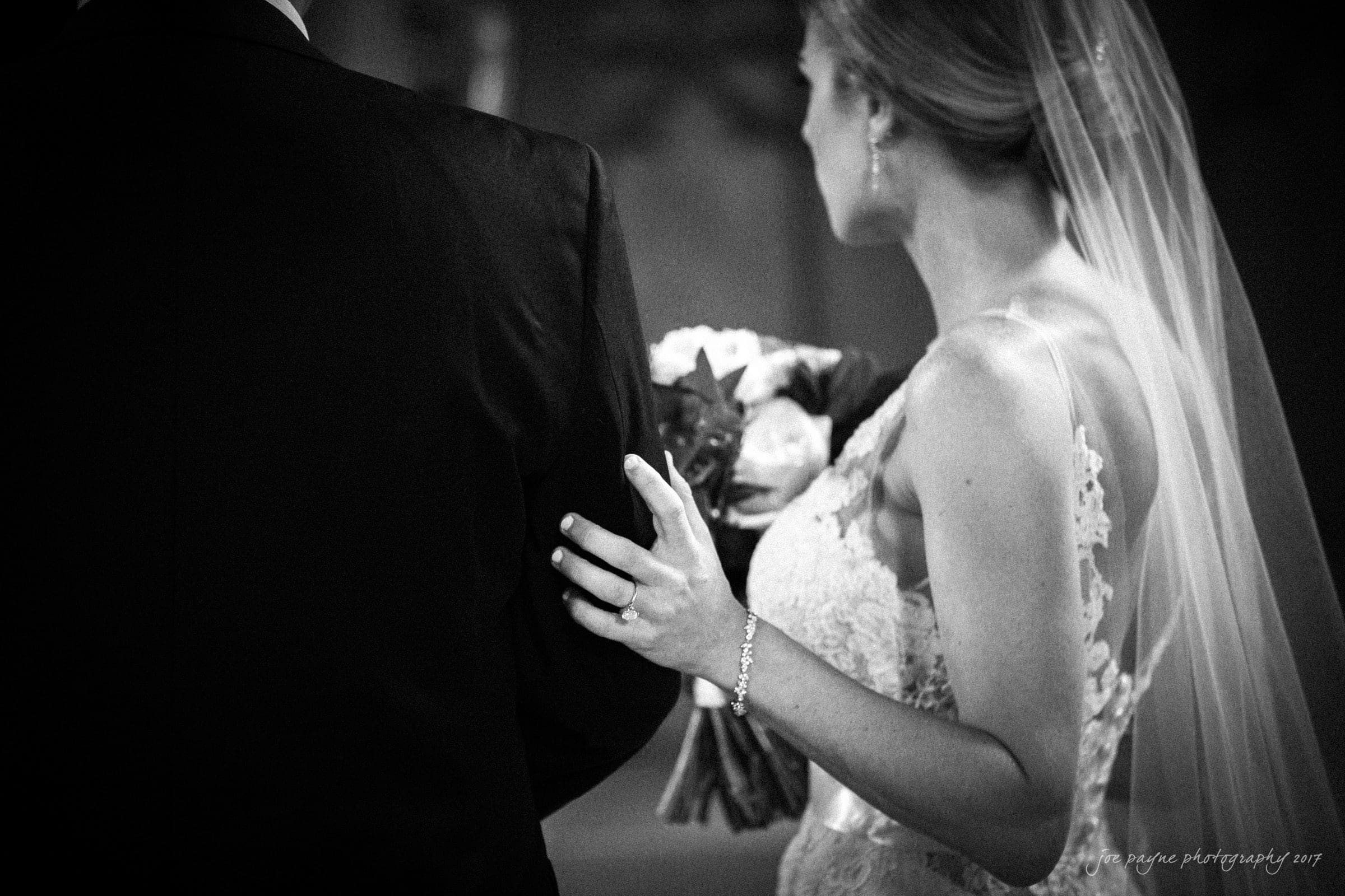 Black and white photo of a bride and groom facing away. The bride, in a lace dress and veil, gently touches the grooms back with her hand, showcasing her ring and bracelet. She holds a bouquet in her other hand.