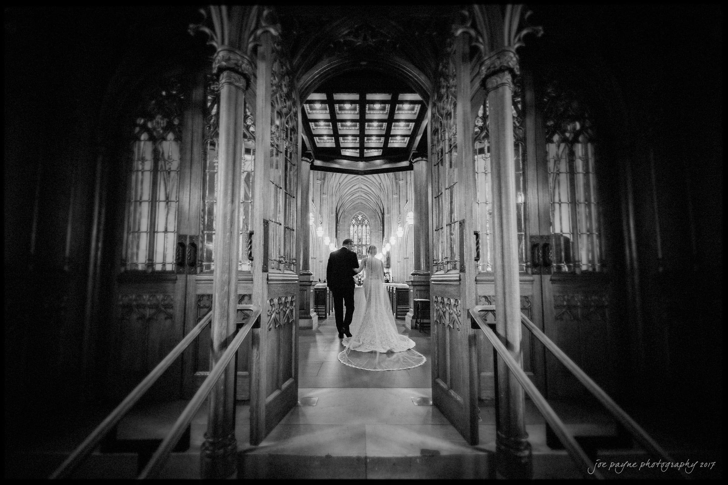 A bride and groom walk hand in hand down an aisle inside a grand, gothic cathedral. The scene is black and white, with intricate architectural details and arched doorways enhancing the romantic atmosphere.