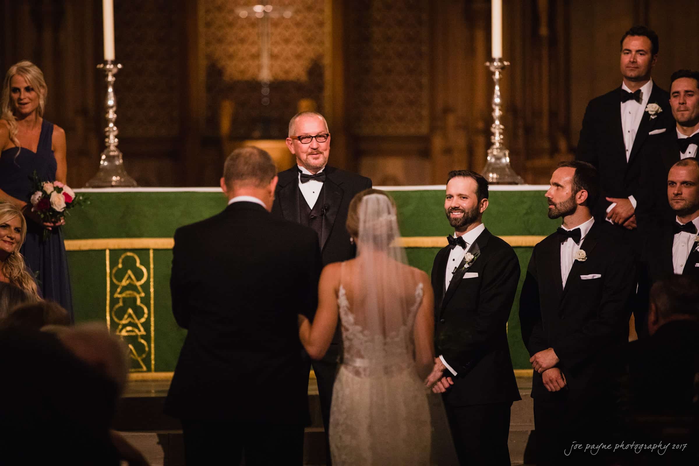 A bride and groom stand before a officiant in a church, surrounded by their wedding party. The groom and groomsmen are in tuxedos, and the bride wears a white gown with a veil. Candles and a green altar cloth are visible in the background.