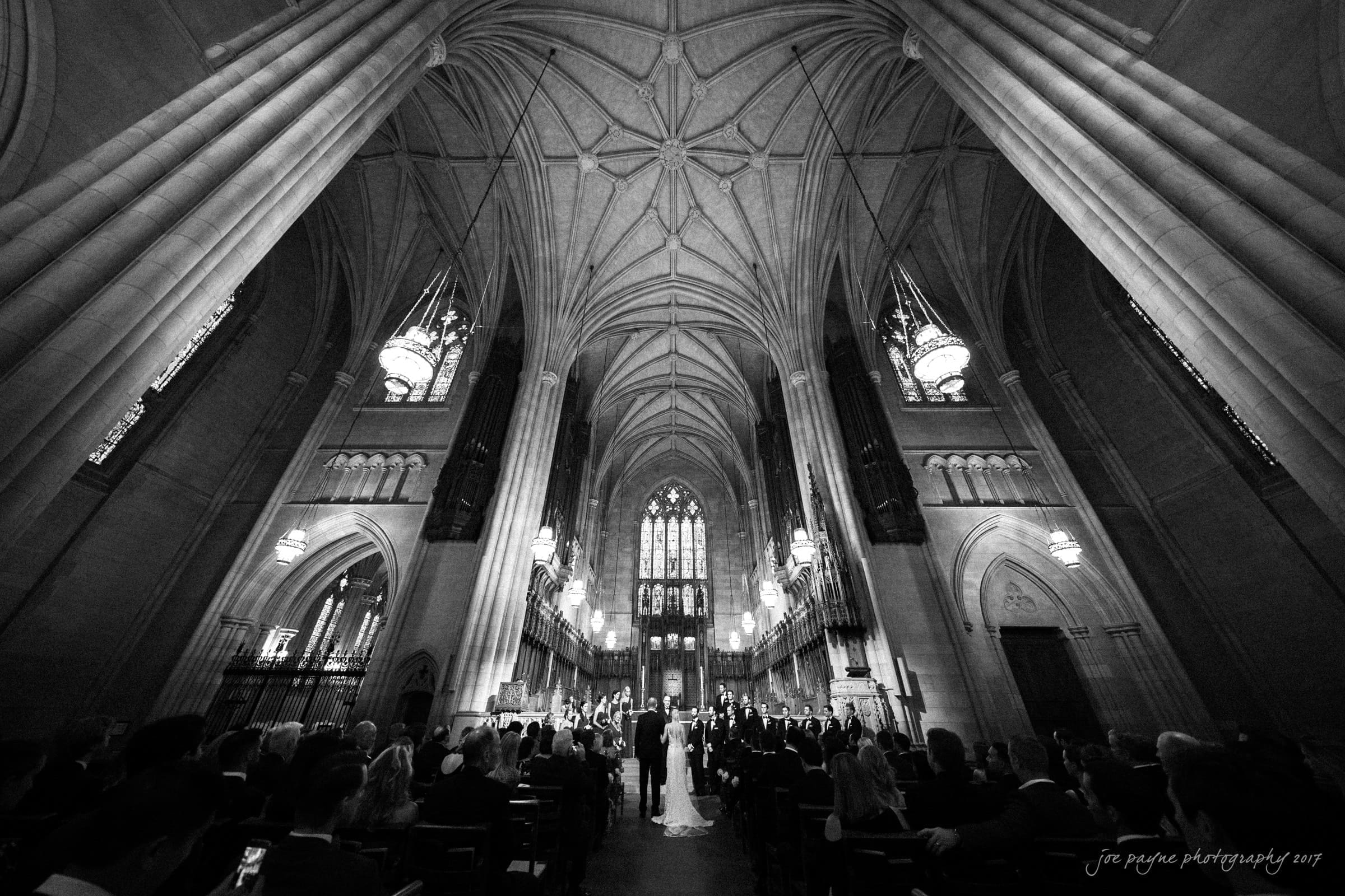 A black and white photo of a cathedral interior during a wedding ceremony. The ornate vaulted ceiling towers above rows of seated guests. The wedding couple stands at the altar, framed by stained glass windows.