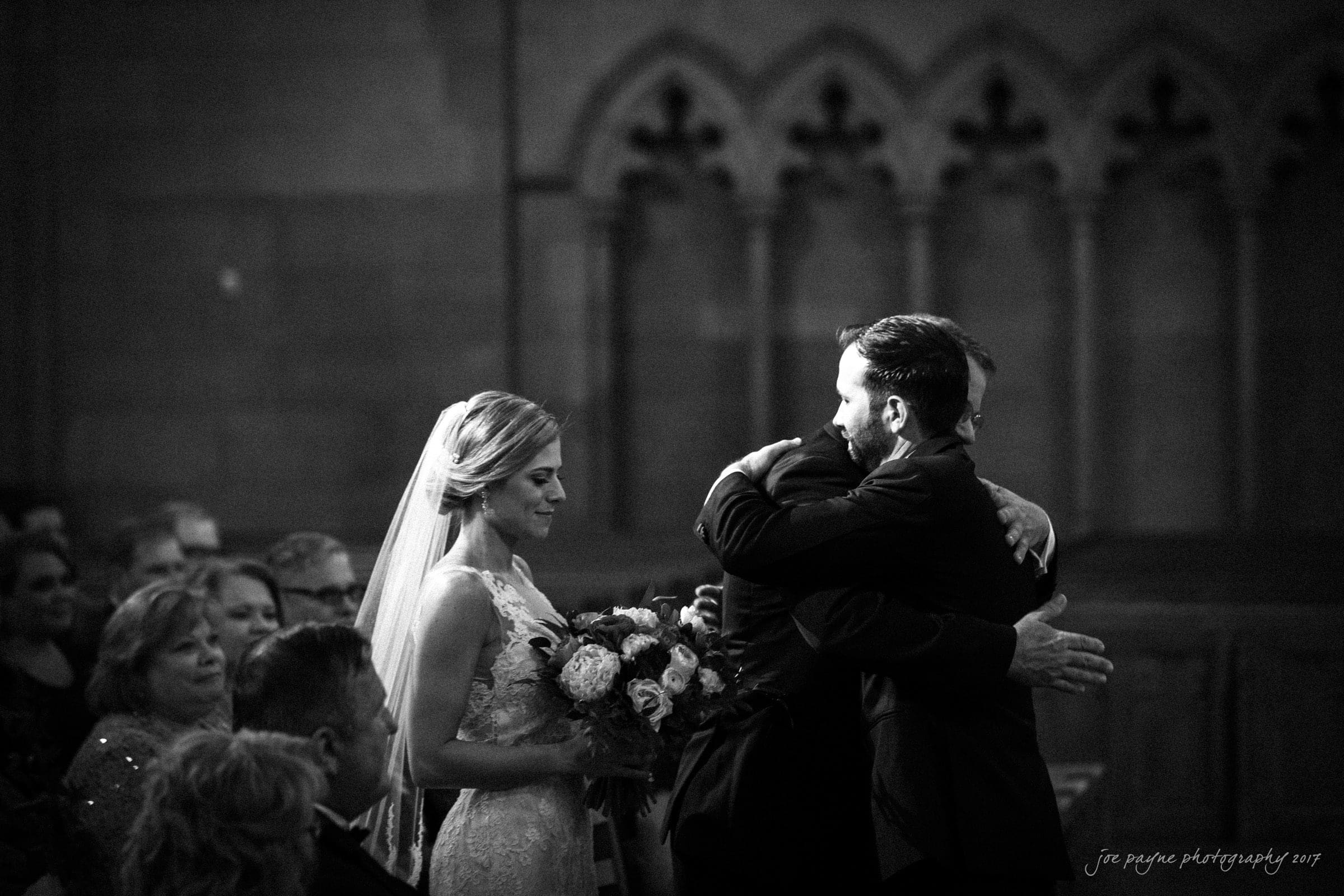 A bride in a wedding dress holds a bouquet and smiles as two men in suits hug each other warmly in a dimly lit church with ornate arches in the background.