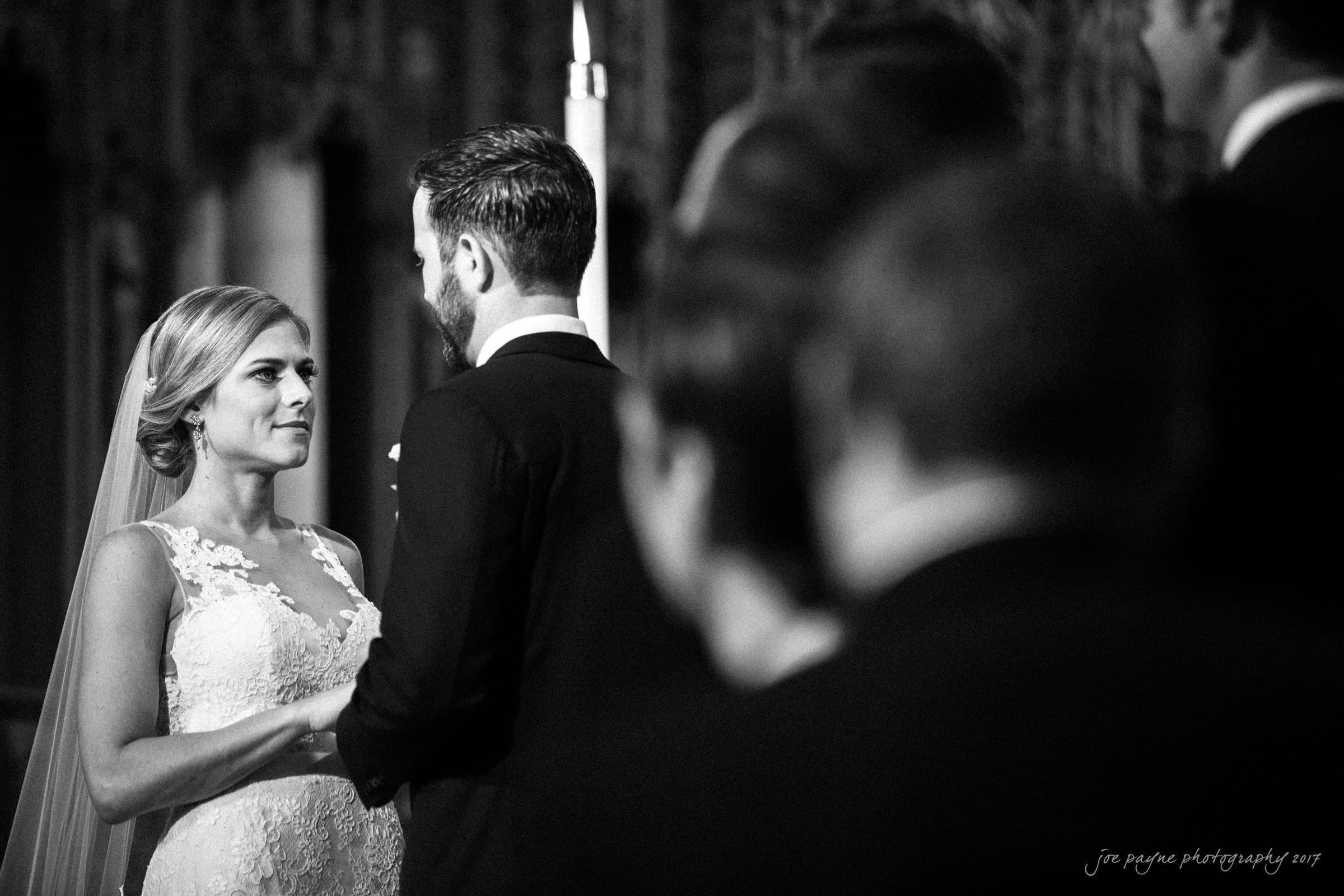 A bride and groom stand facing each other, holding hands. The bride wears a lace dress and veil, while the groom is in a suit. The scene appears emotional and intimate, hinting at a wedding ceremony. Other people are blurred in the foreground.