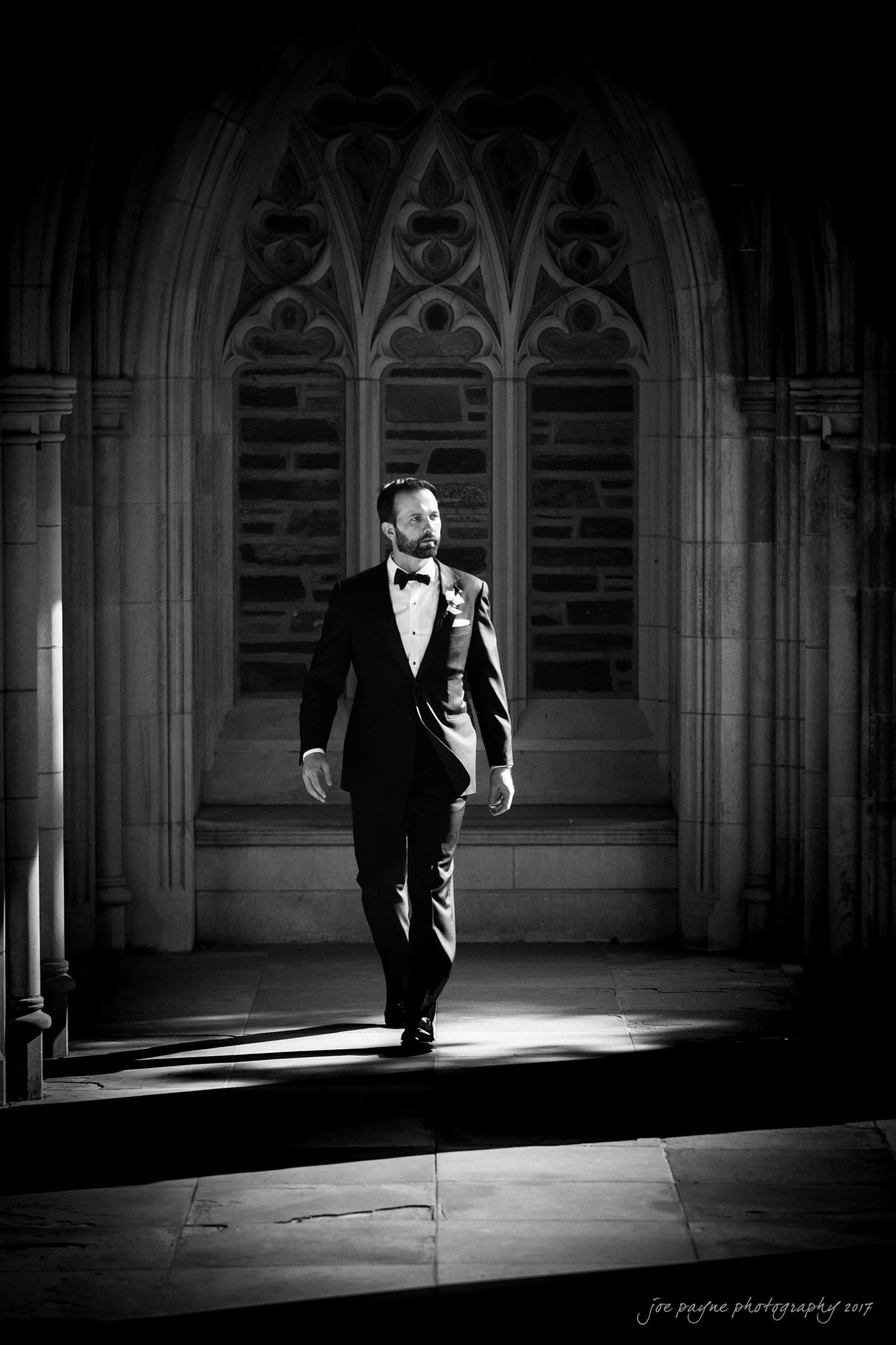 A man in a tuxedo walks confidently down a sunlit path, framed by ornate arches. The setting is a dramatic, gothic architectural environment with detailed stonework.