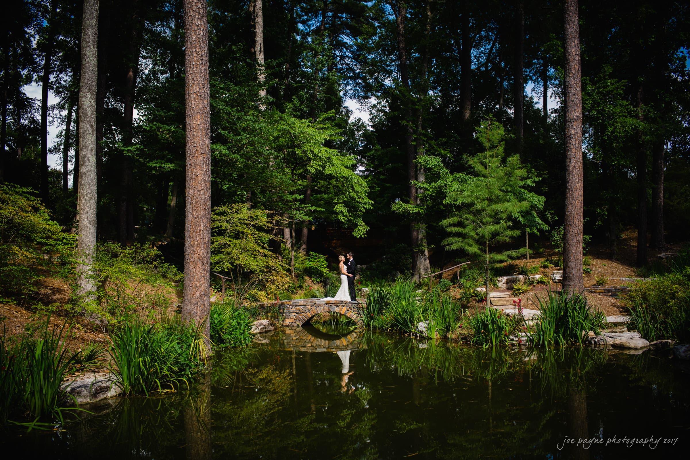 A couple stands on a small stone bridge over a tranquil pond in a lush, forested garden. Tall trees and vibrant greenery surround them, with their reflection visible in the water.