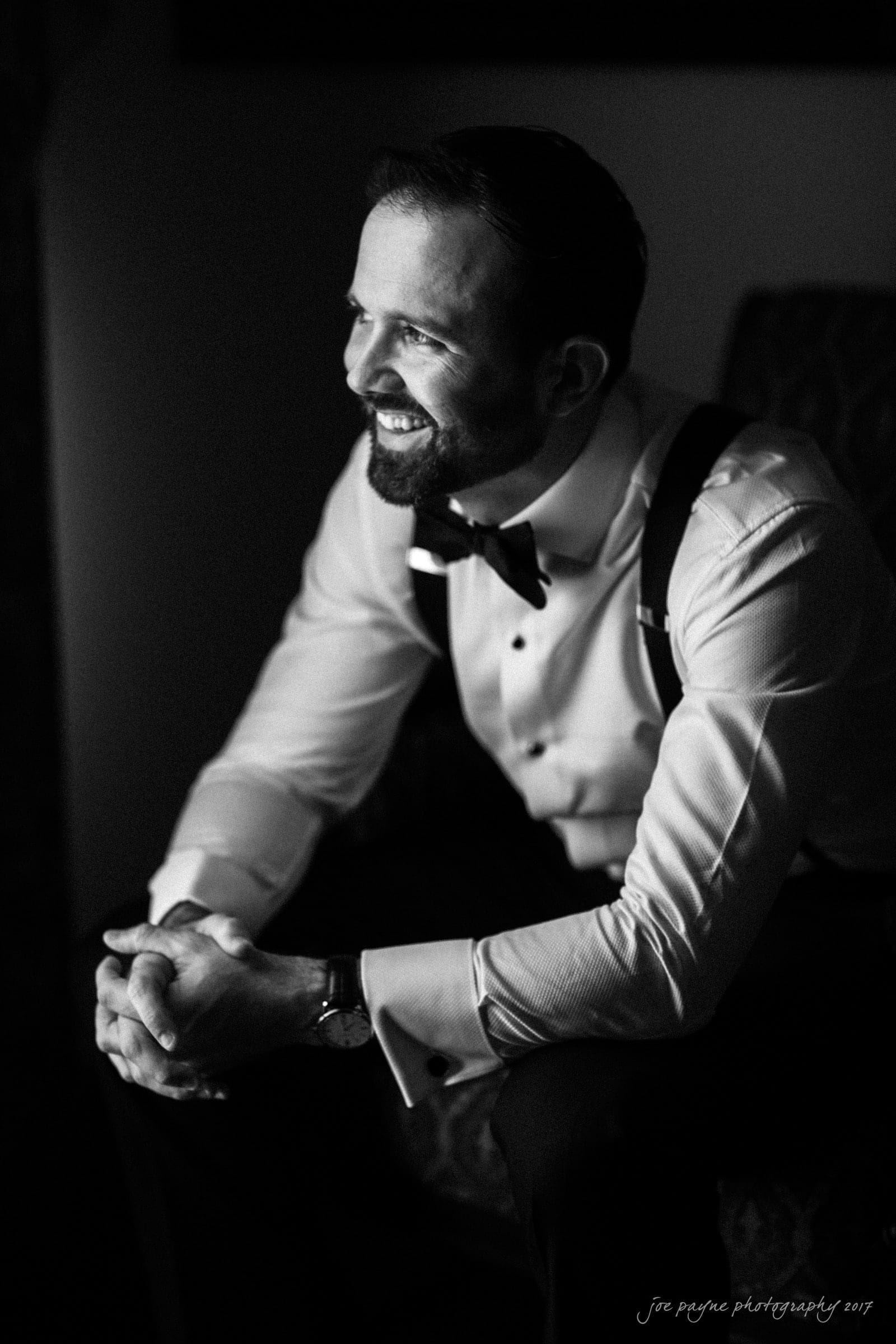 Black and white photo of a man sitting in a chair. He is wearing a dress shirt, bow tie, and suspenders. He is smiling and looking to the side, hands clasped in front of him. Soft lighting creates a contrast, highlighting his features.