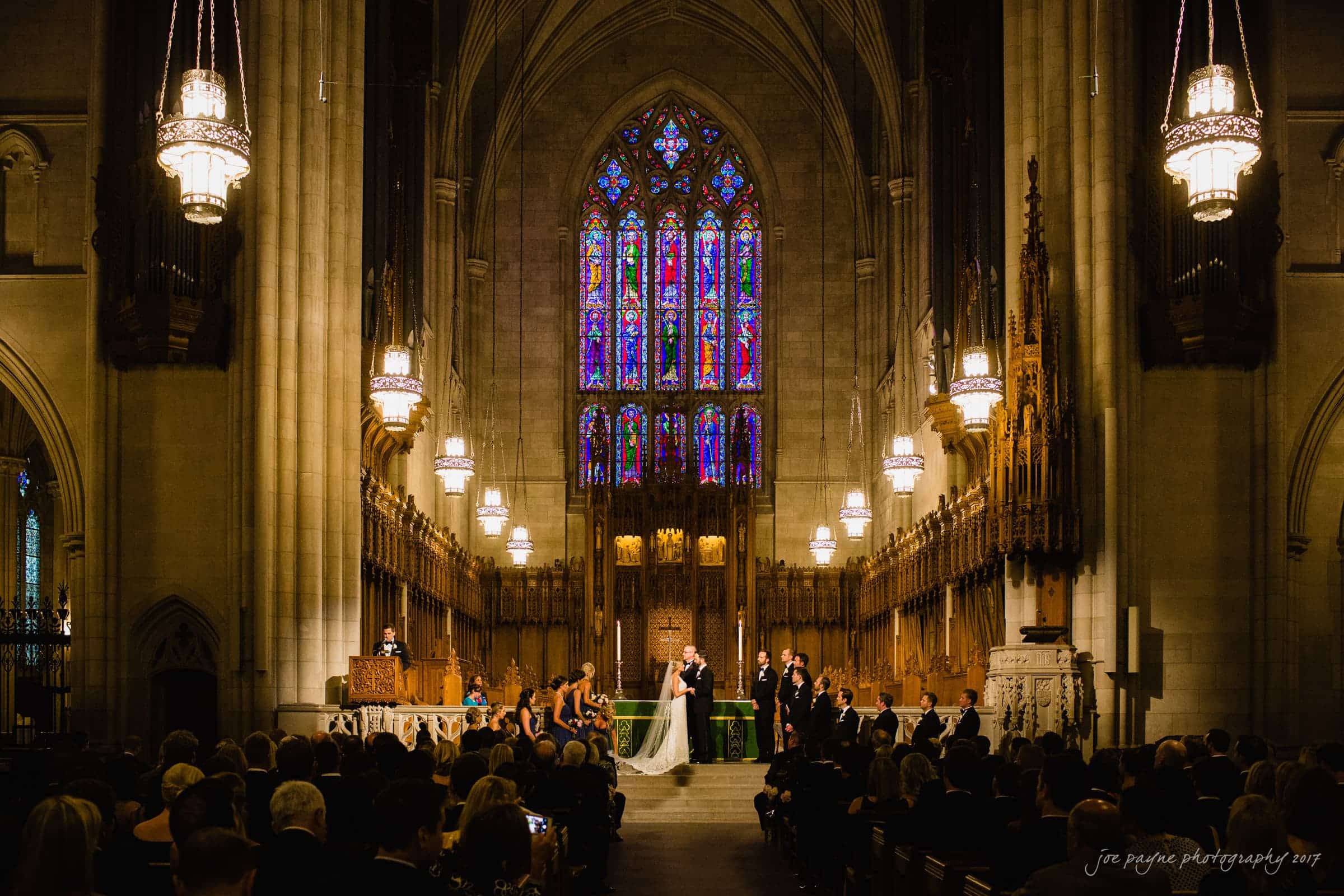 A grand church interior during a wedding ceremony. The bride and groom stand at the altar, surrounded by bridesmaids and groomsmen. Stained glass windows and ornate chandeliers illuminate the scene. Guests are seated, watching the ceremony.