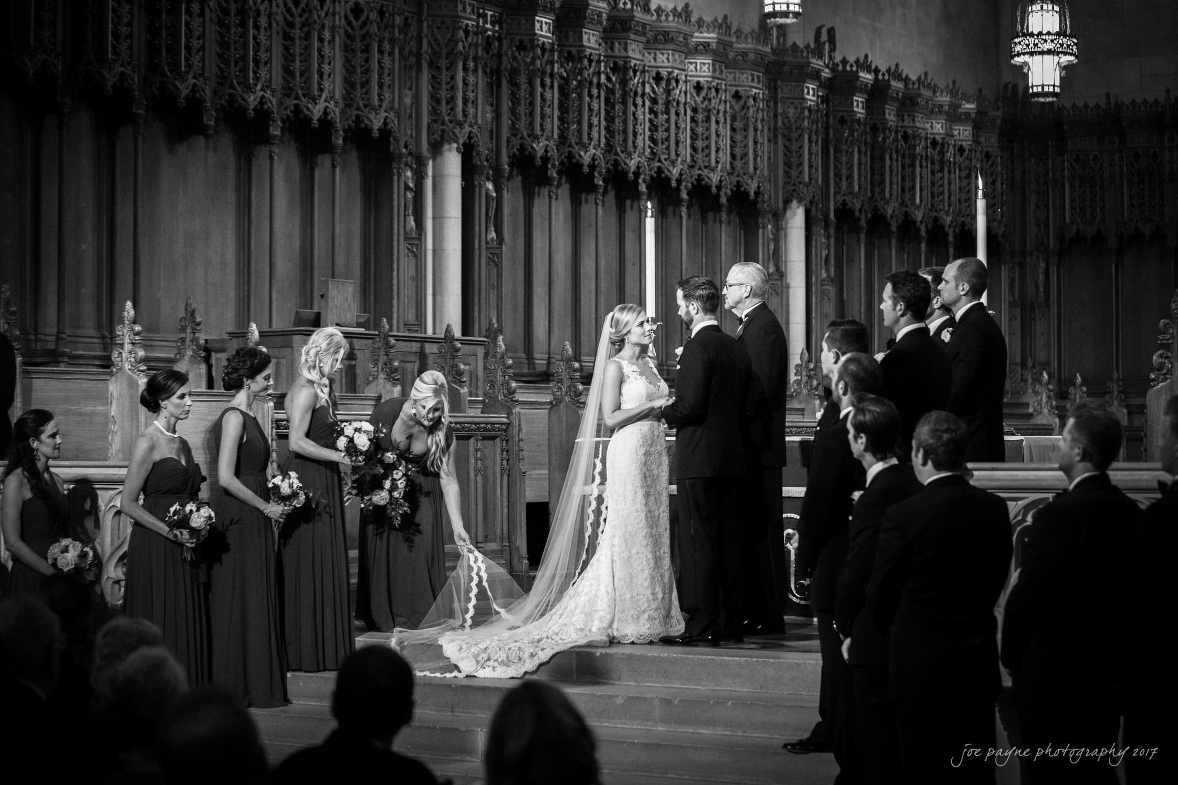 A black and white photo of a wedding ceremony in a grand church. The bride and groom stand at the altar with the officiant. Bridesmaids in matching dresses hold bouquets, and groomsmen in suits stand on either side. Guests are seated in the foreground.